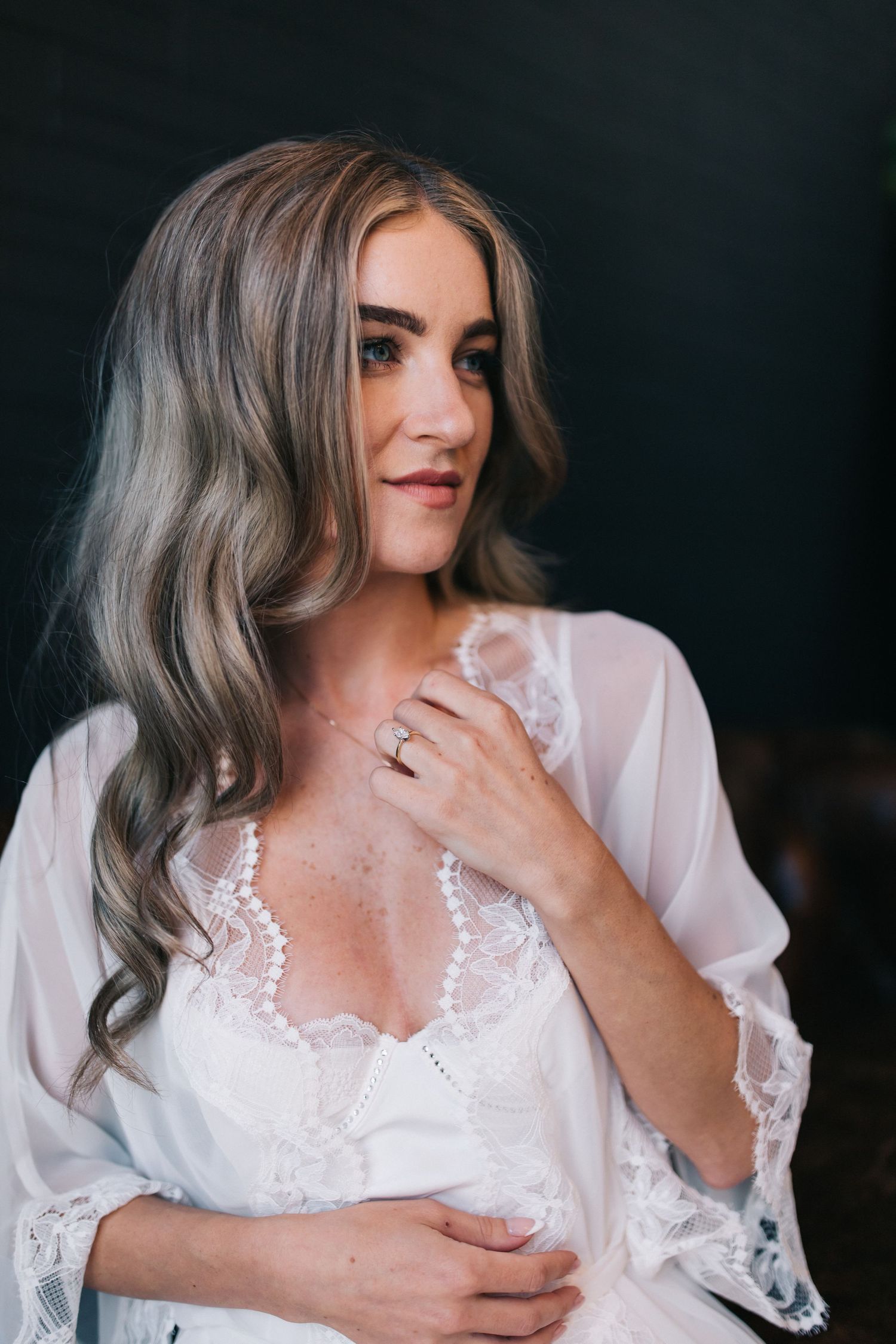 Woman in white lace robe with soft wavy hair poses against dark background.