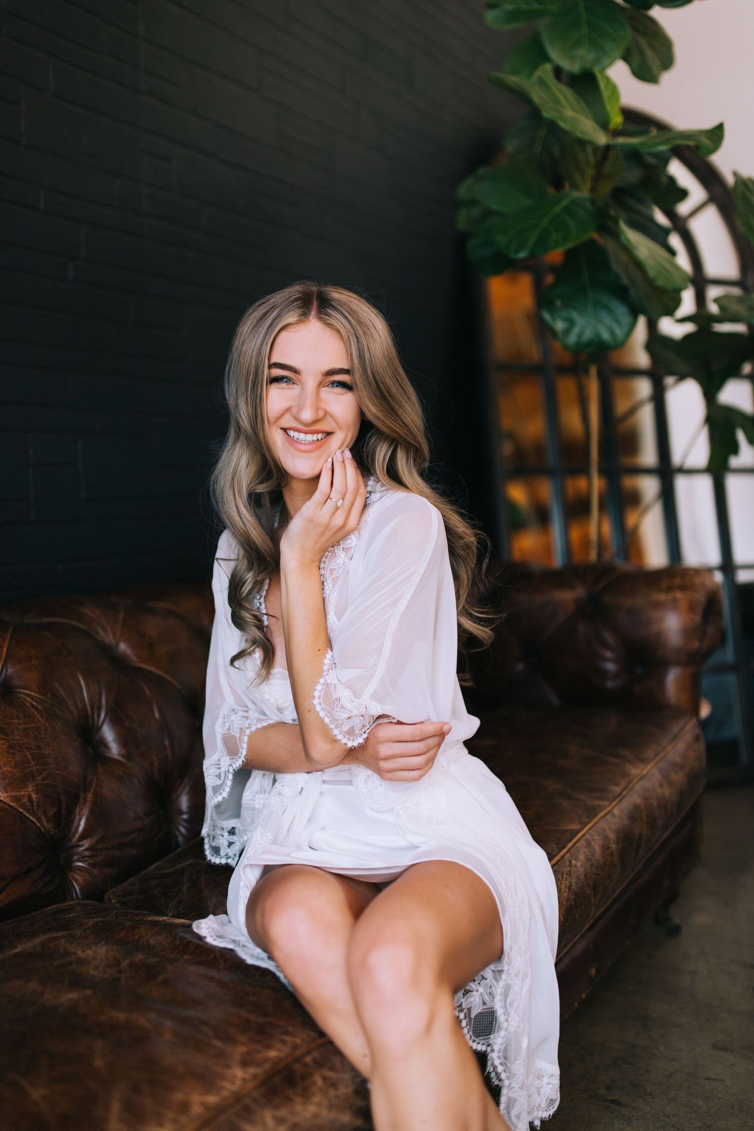 Woman in white lace robe sits smiling on brown leather couch with plants in background.