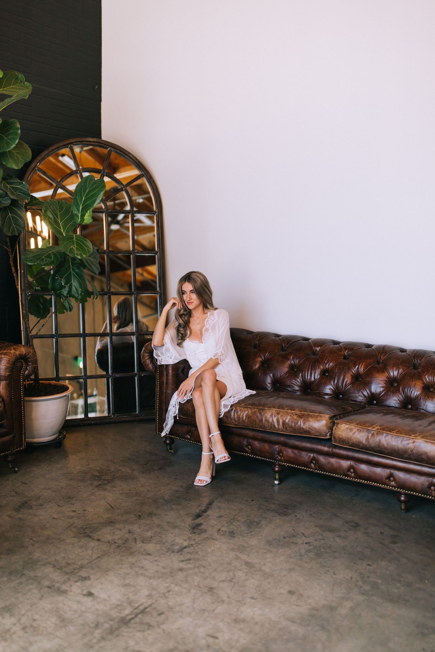 Woman in white robe poses on vintage leather couch near arched mirror and plants.