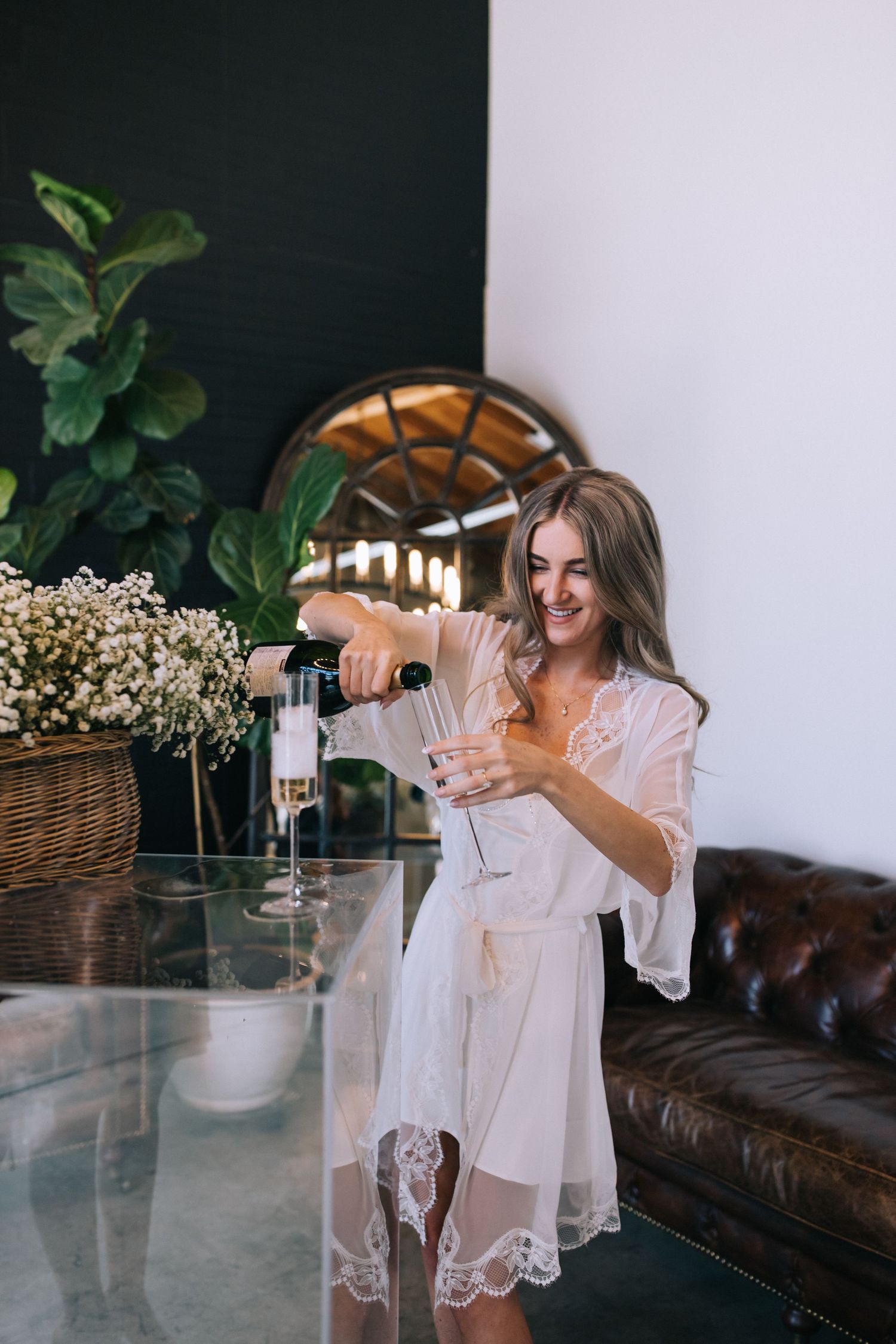 Woman in white lace robe pours champagne at modern table with baby's breath flowers.