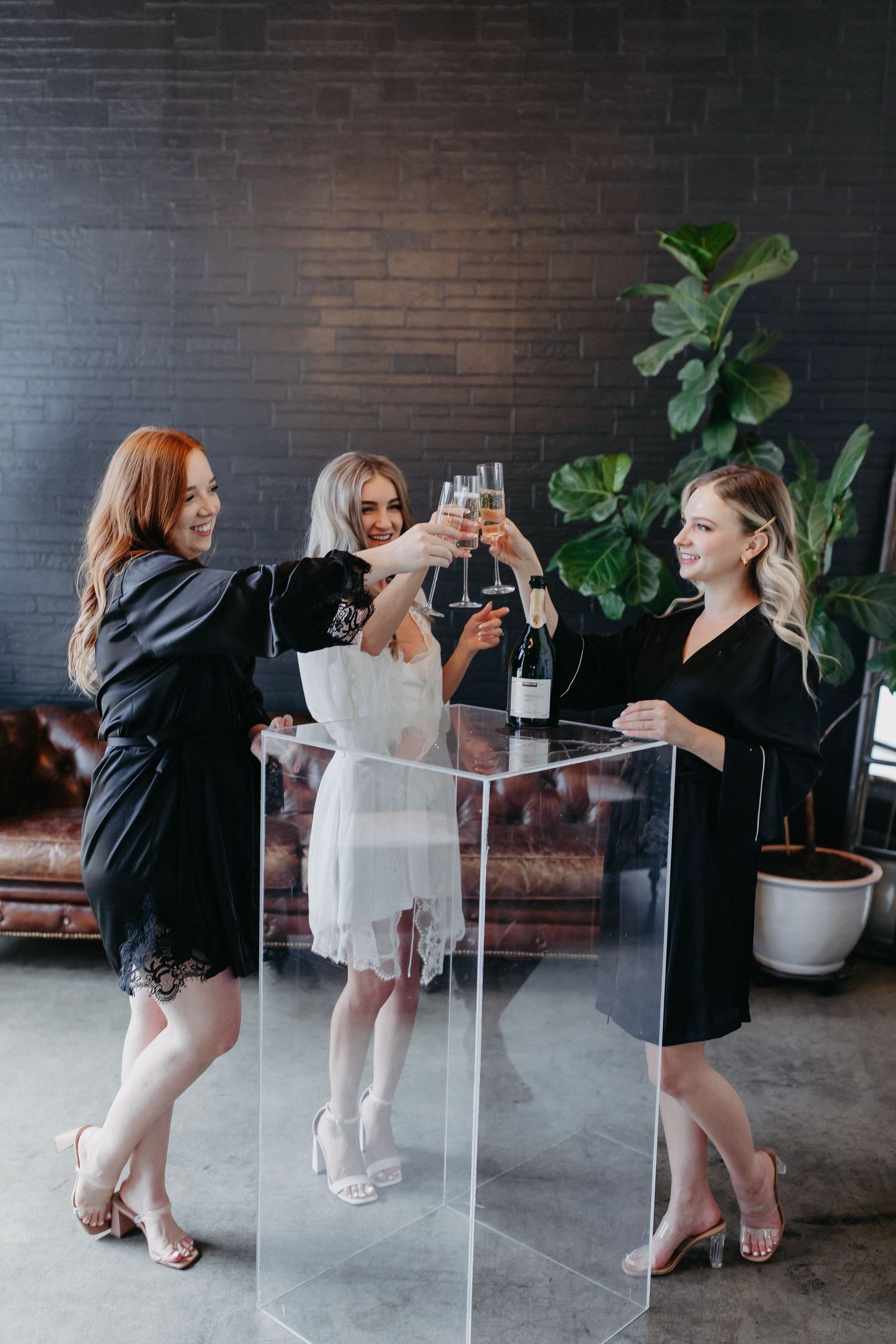 Three women in elegant dresses toast with champagne glasses by leather sofa.