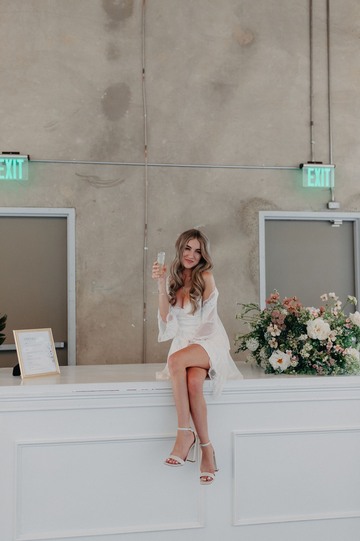 Woman in white dress sitting on white counter near exit signs and floral arrangement.