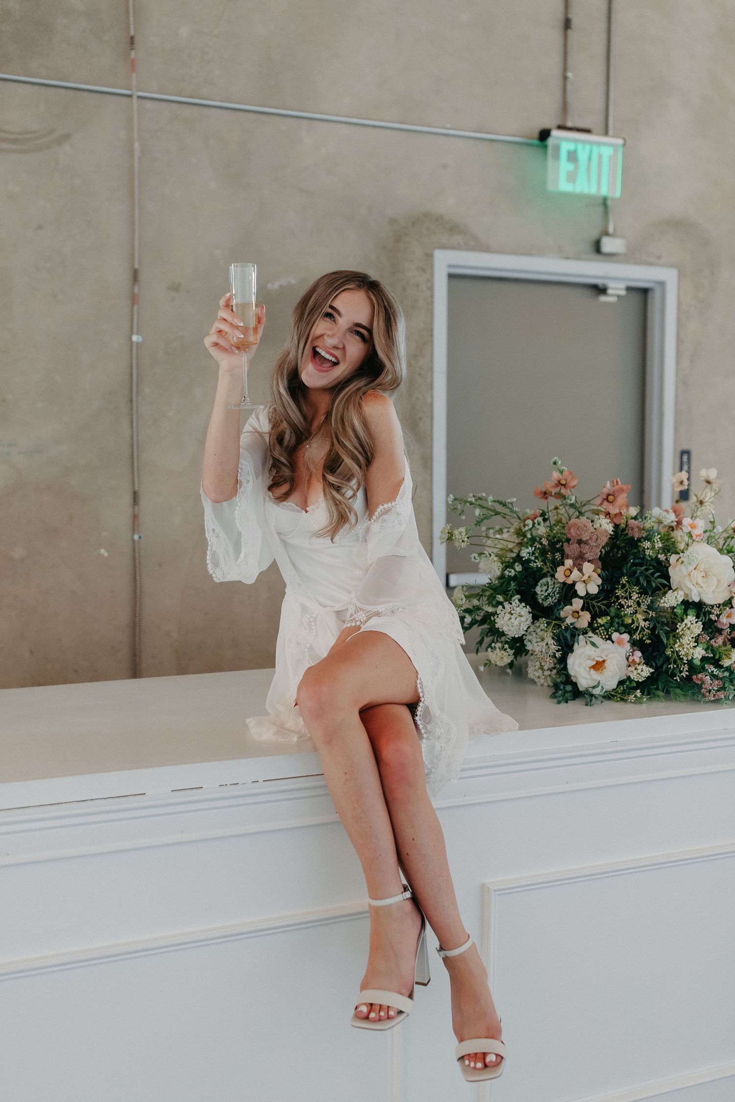 Woman in white dress raising champagne glass while sitting on white ledge with floral decorations.