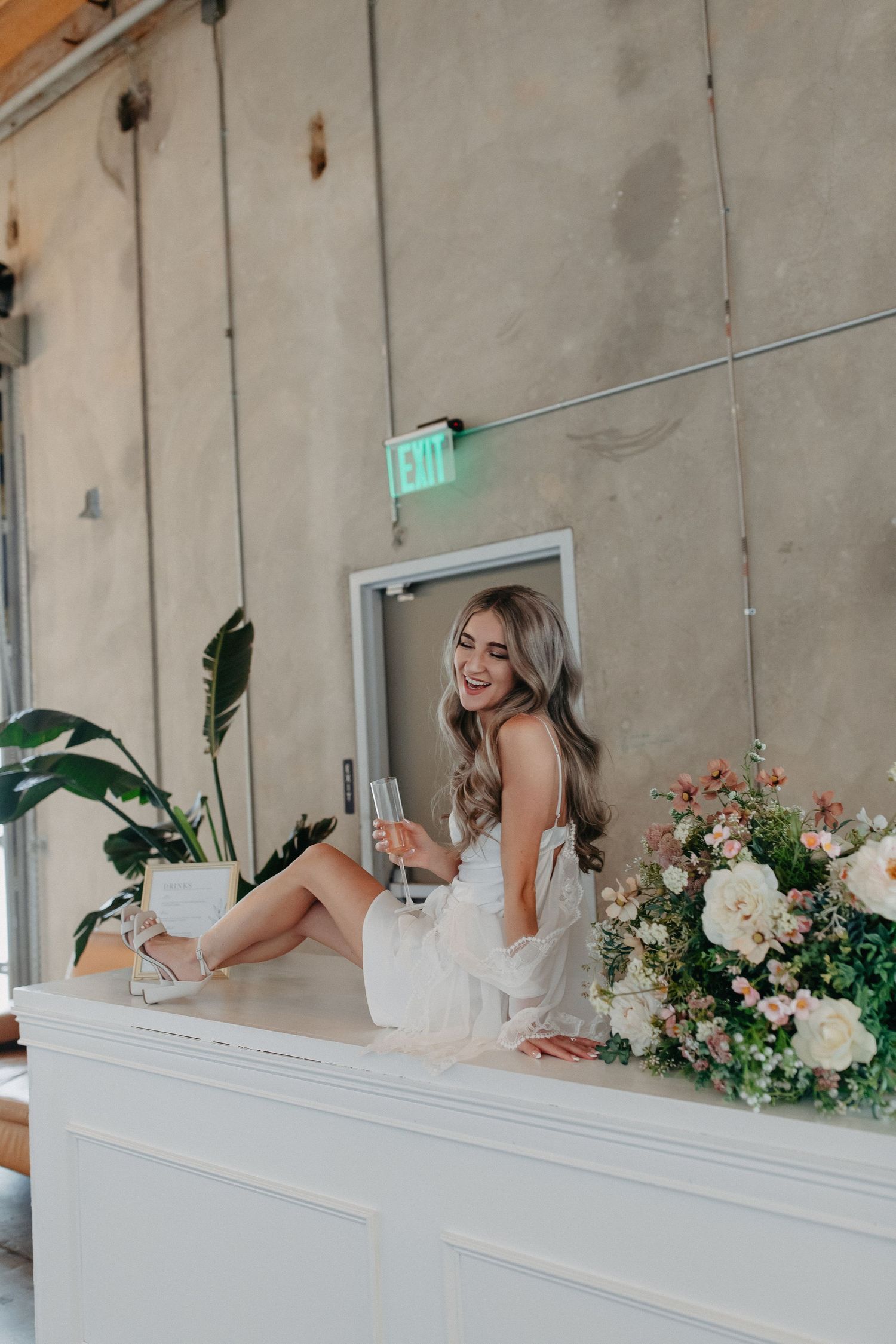 Bride sits on white ledge with glass of champagne and floral arrangements against industrial wall.