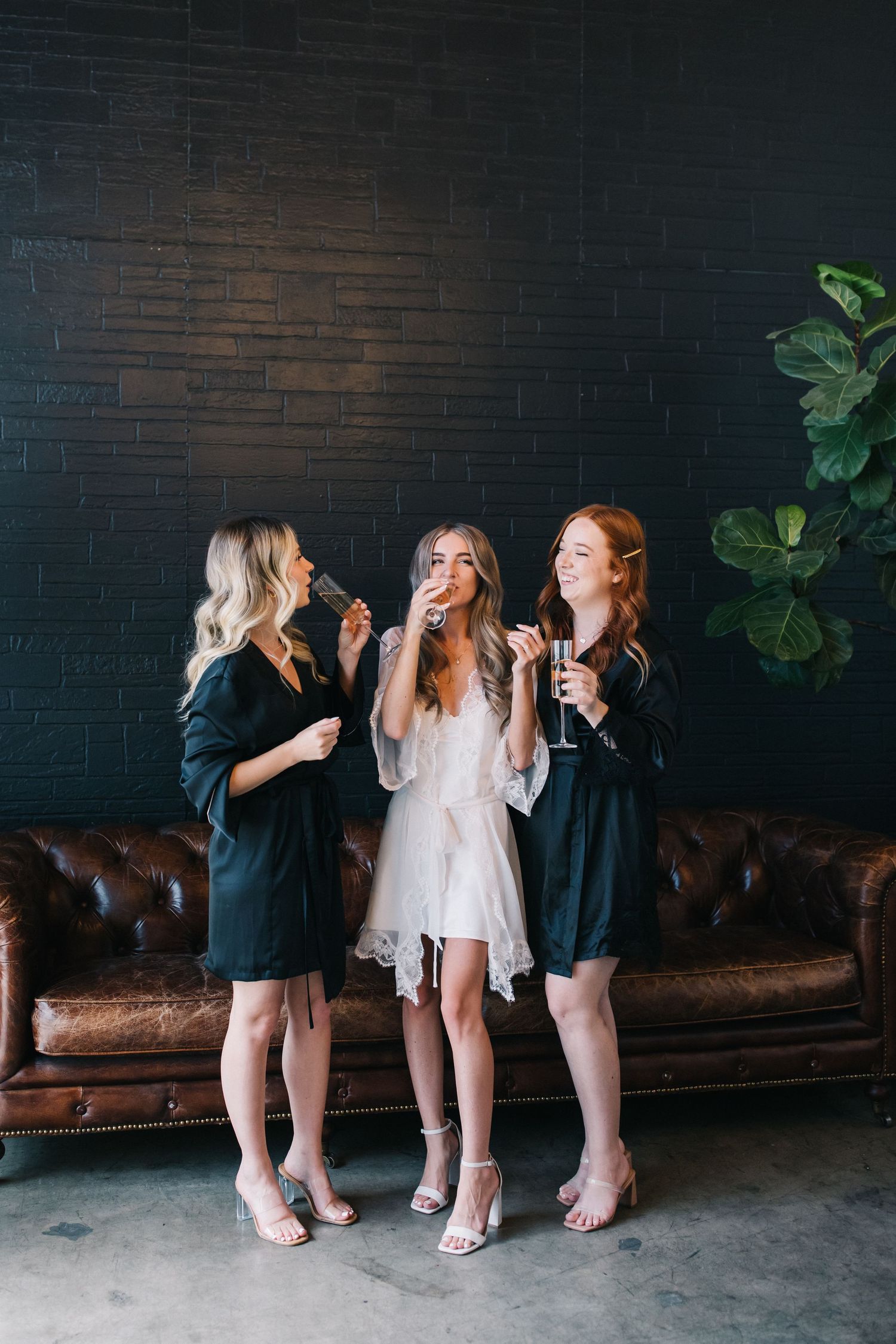 Three women in formal attire enjoying drinks and laughter against dark brick wall with leather couch.