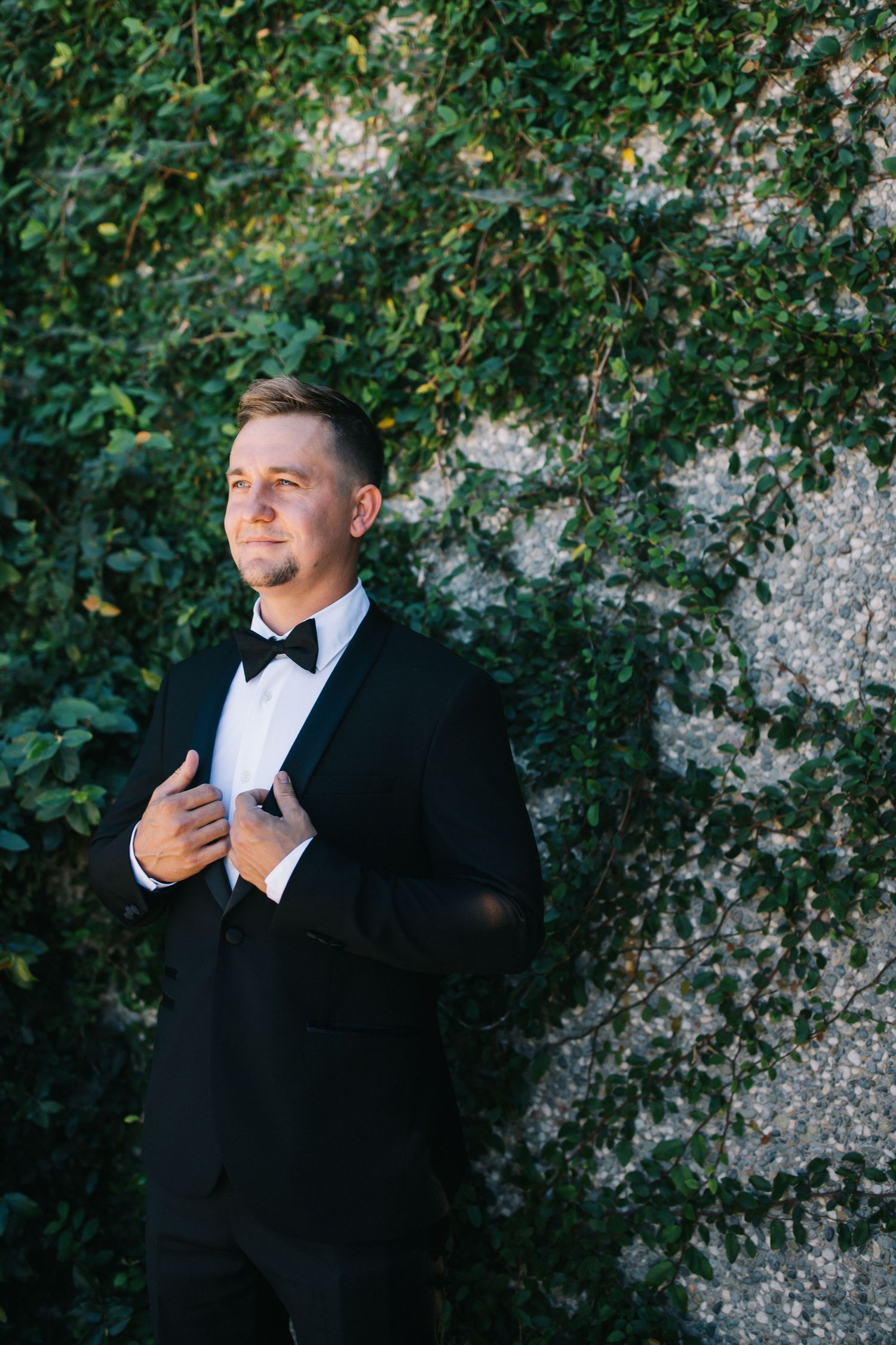 Groom in classic black tuxedo and bowtie poses against leafy garden wall.