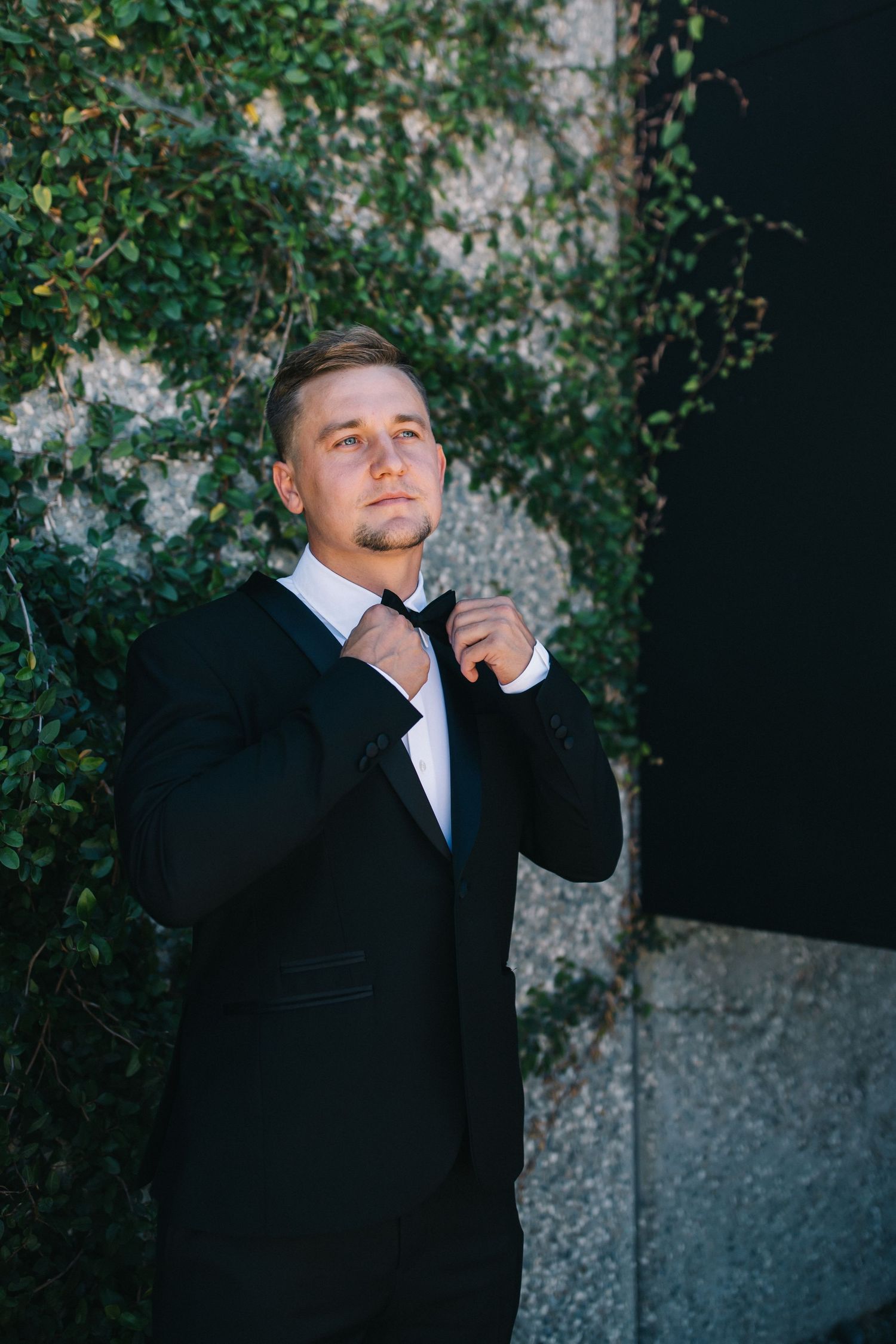 Formal portrait of groom adjusting tie against ivy wall background.