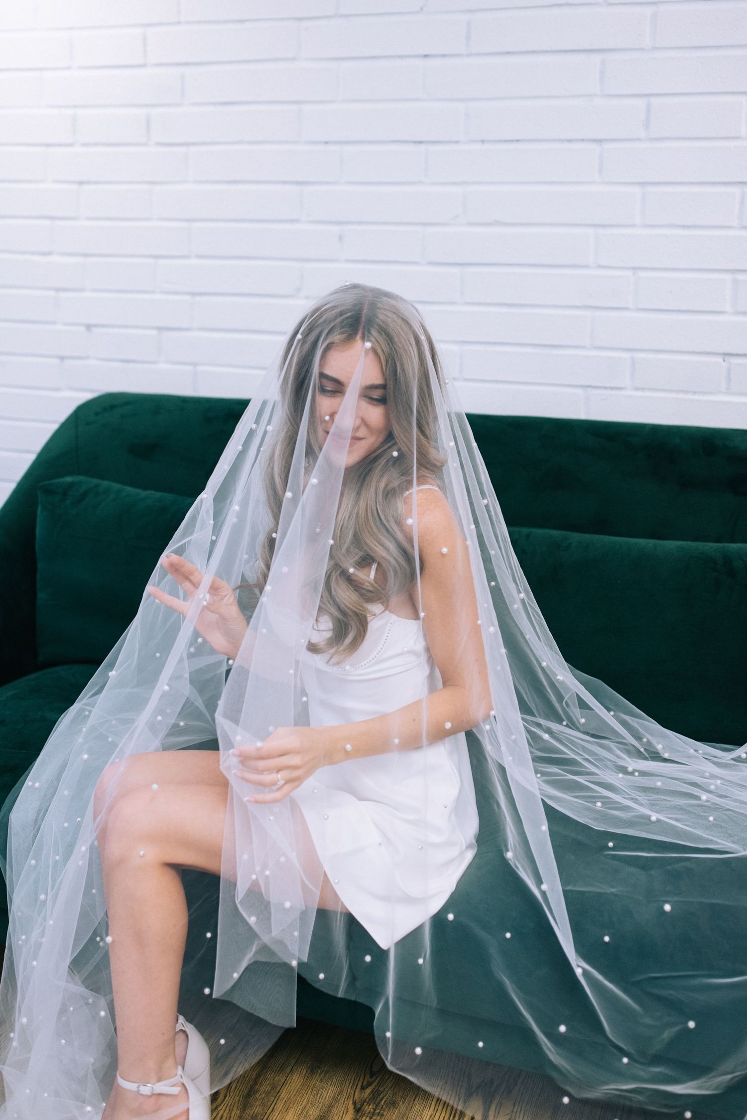 Woman in white dress sitting on green velvet couch covered in flowing bridal veil.