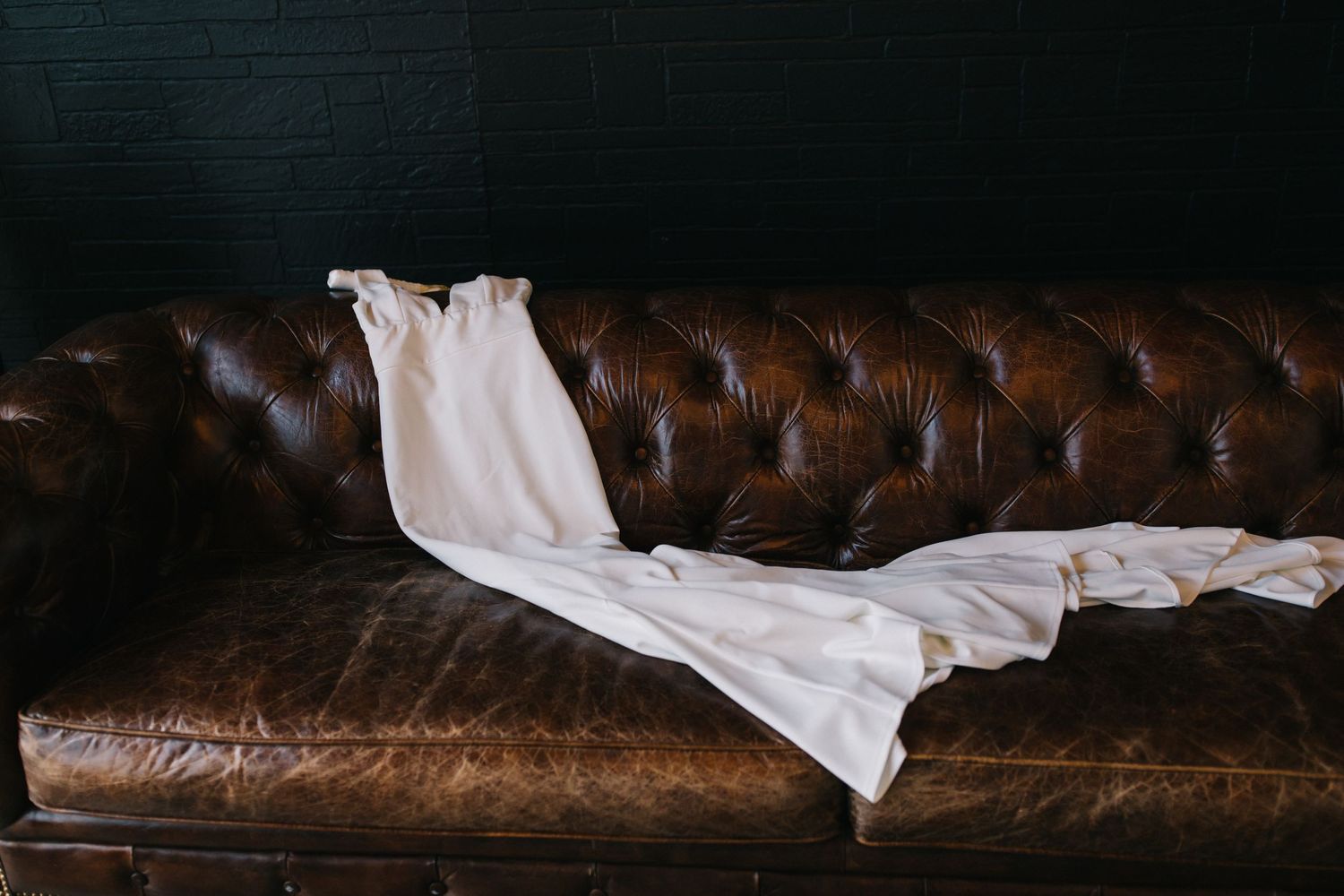 White wedding dress draped over vintage brown leather couch in minimalist setting.