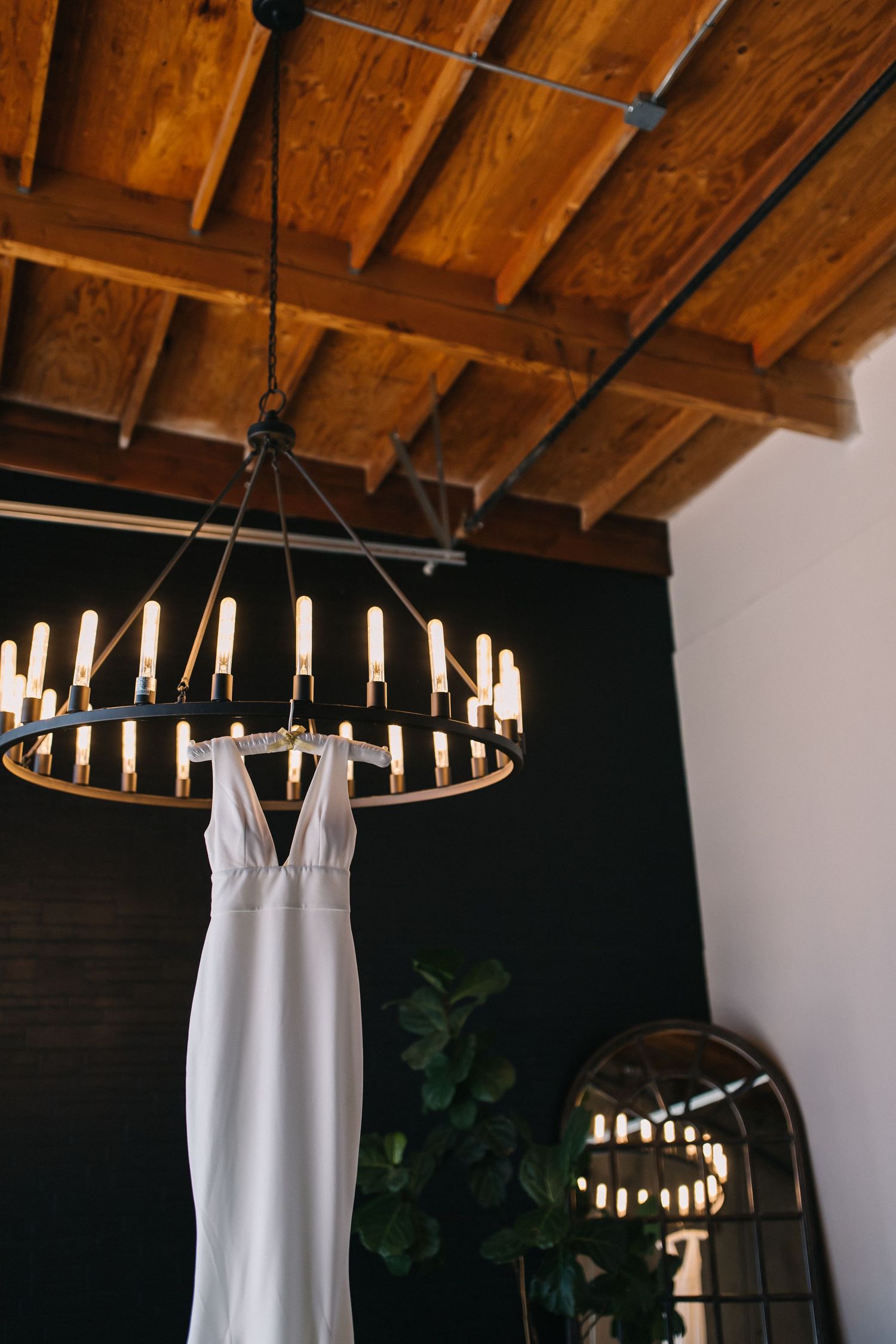Elegant white wedding dress hanging from circular chandelier against dark wall with wooden beams.