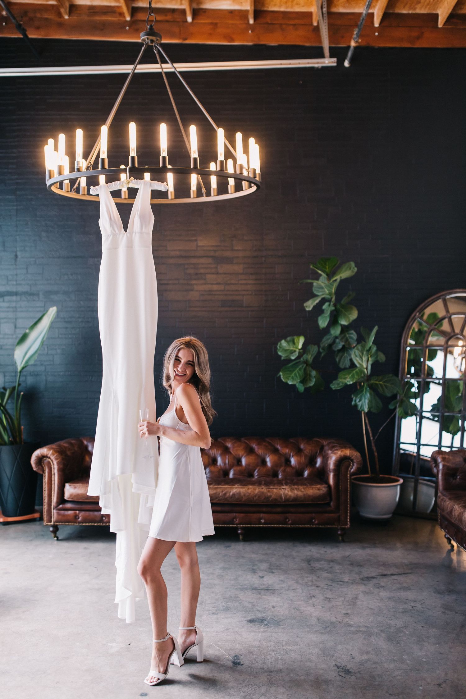 Woman in white dress standing next to hanging wedding gown under circular chandelier.