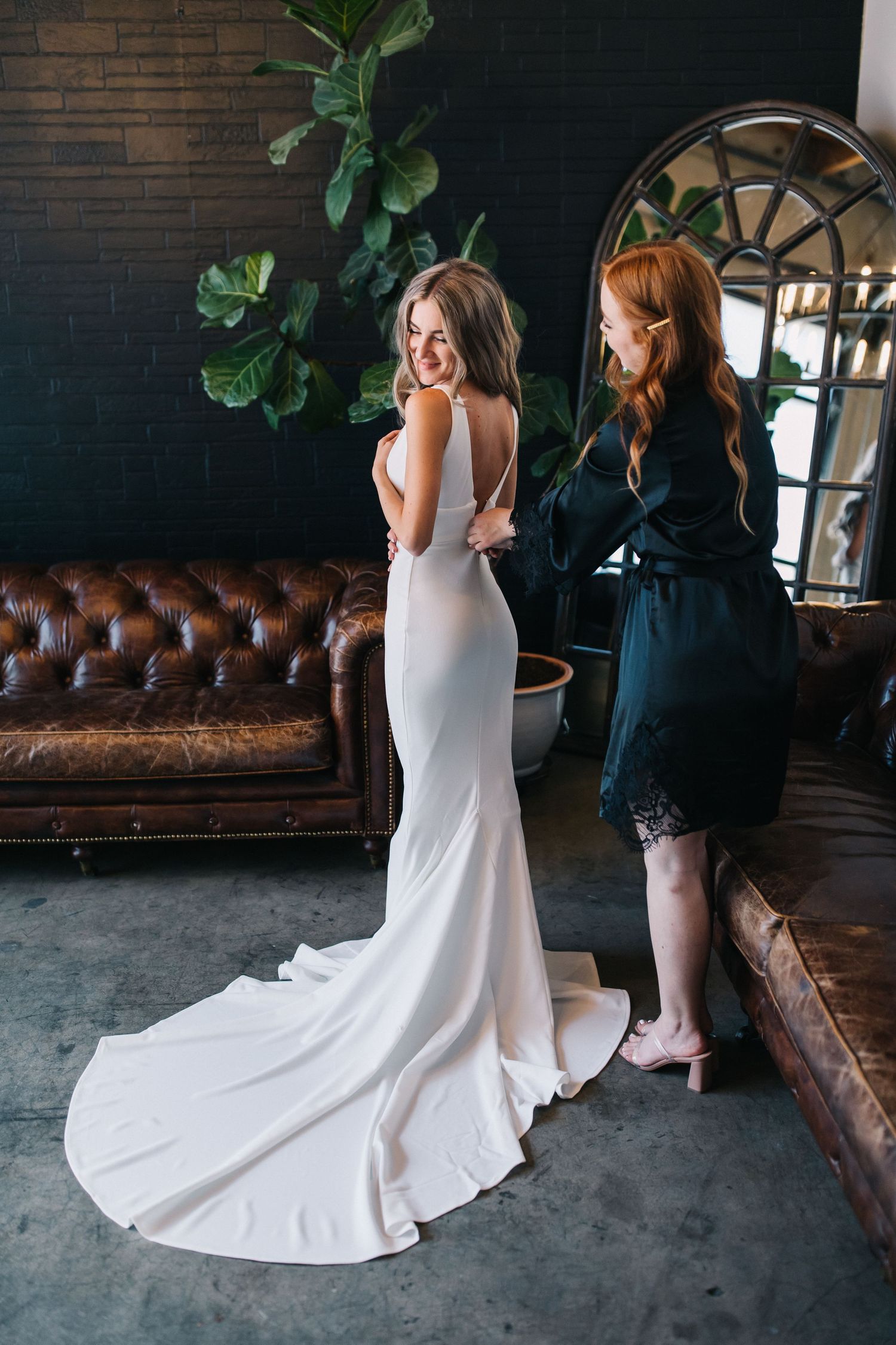 Woman in flowing white wedding gown being helped with dress adjustments near leather couch.