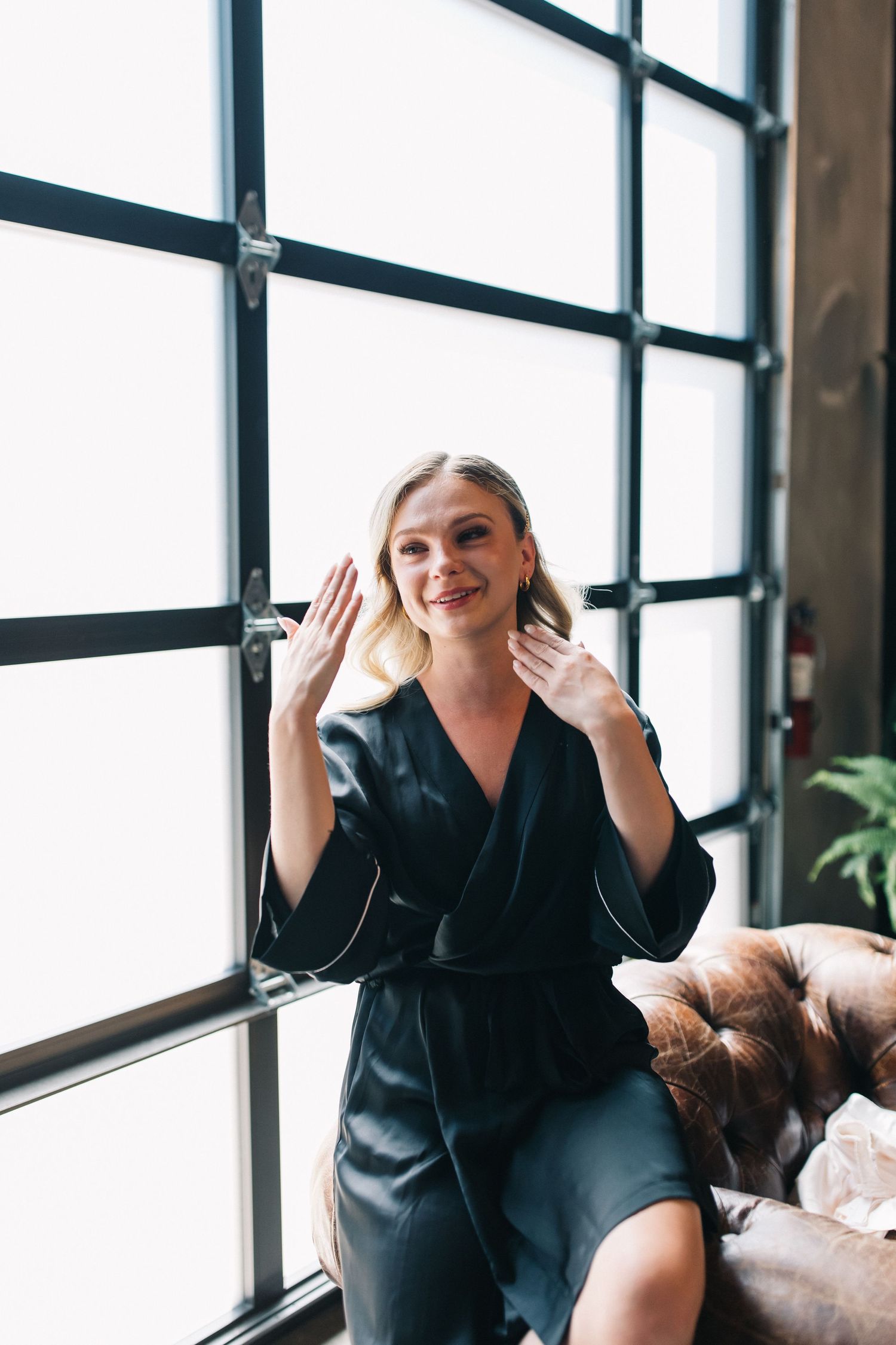 A person in a black dress sits by a large window waving with a cheerful expression.