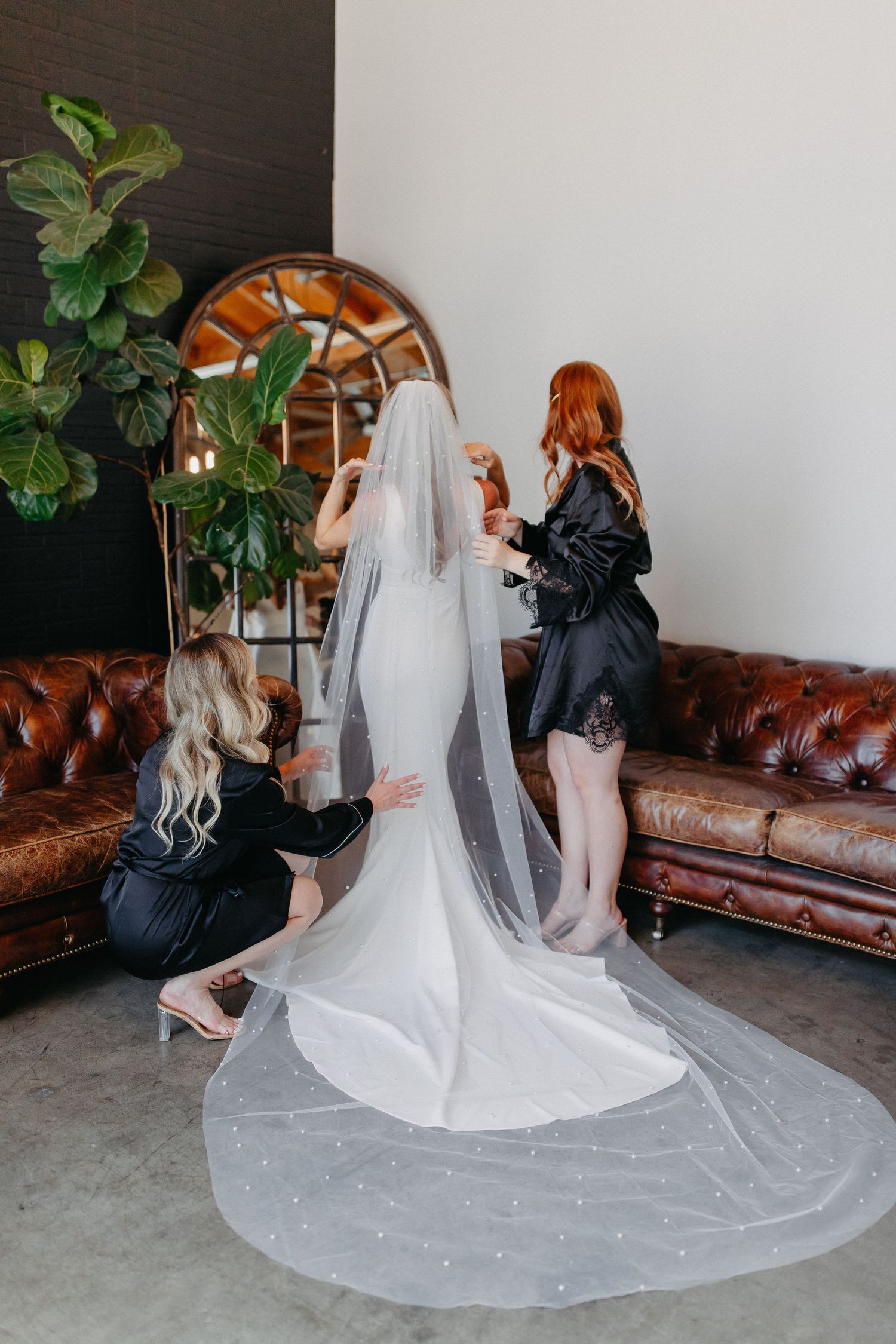 A bride in a fitted white gown gets her veil adjusted near a decorative arched mirror.
