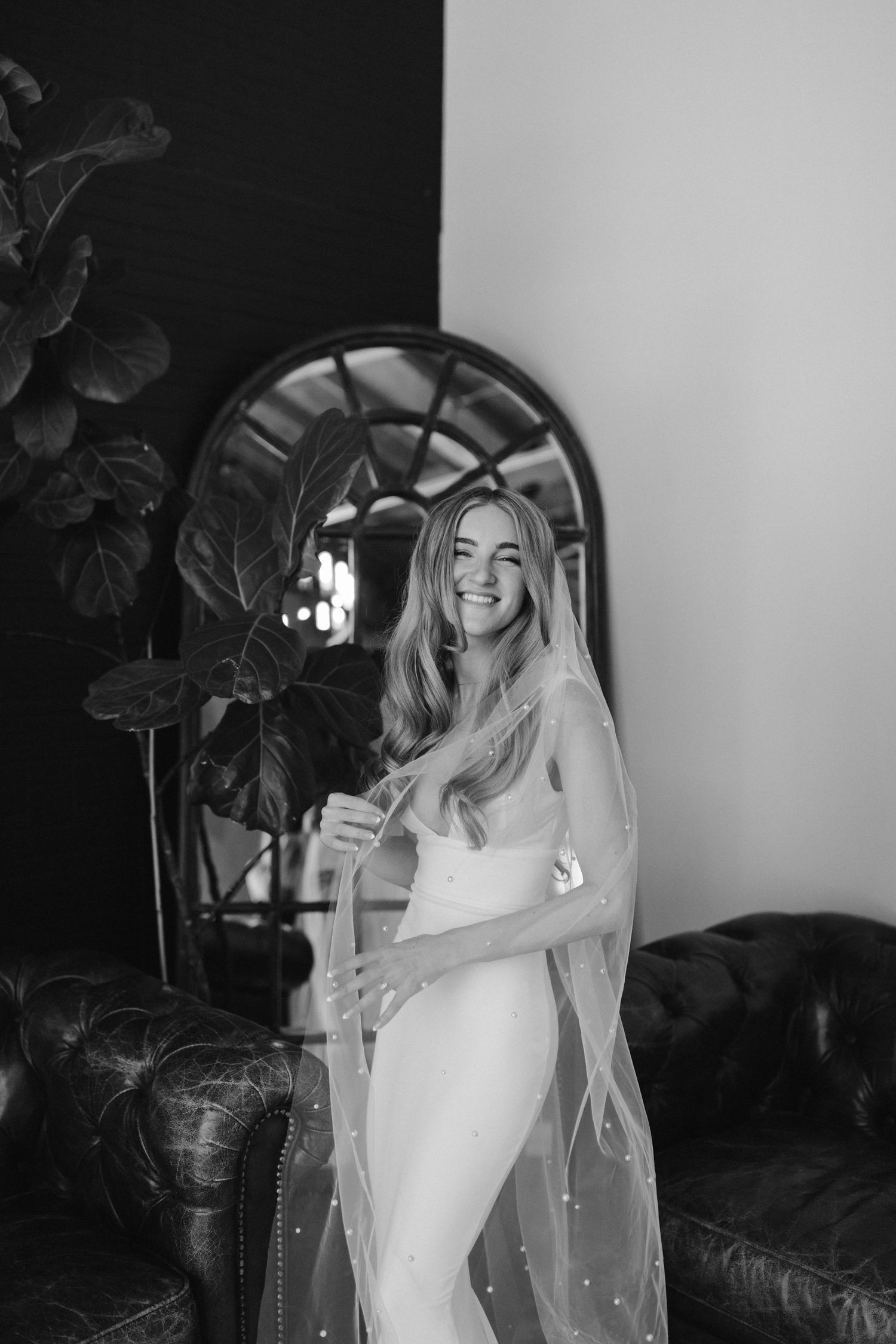 A bride poses in an elegant white fitted wedding dress with a long veil next to a vintage leather couch and arched mirror.