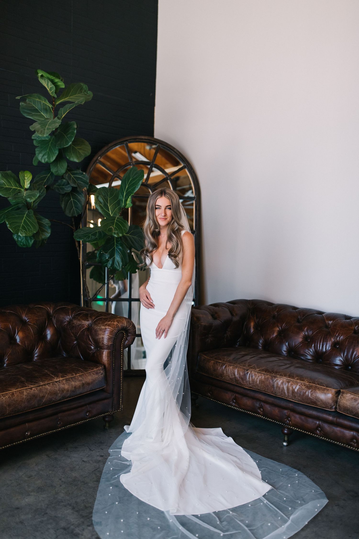 A bridal portrait featuring a fitted white gown with long train in a room with leather couches and fiddle leaf fig plant.