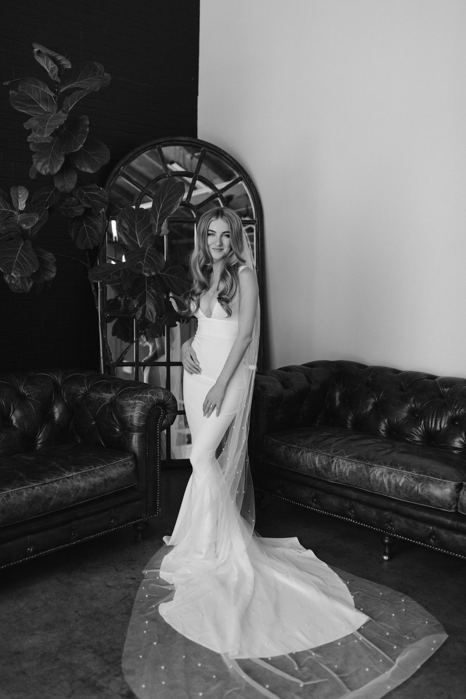 Black and white photo of a bride in a sleek wedding dress standing between leather sofas with an arched mirror.