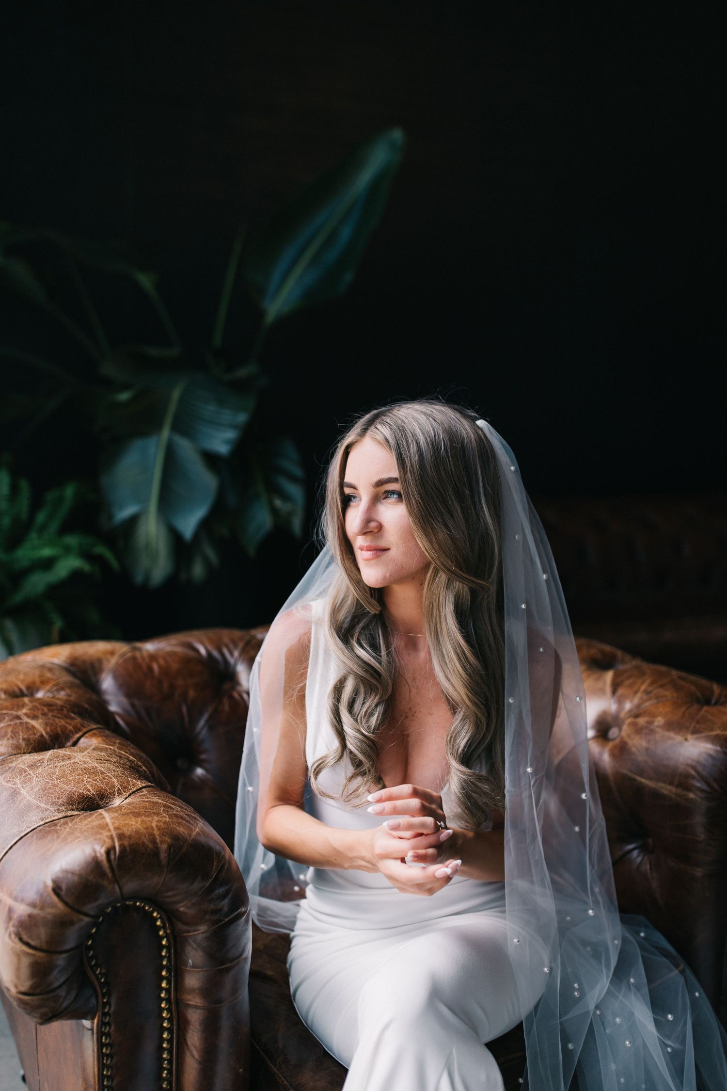 Candid bridal portrait in a white wedding dress and veil seated on a brown leather chesterfield sofa.
