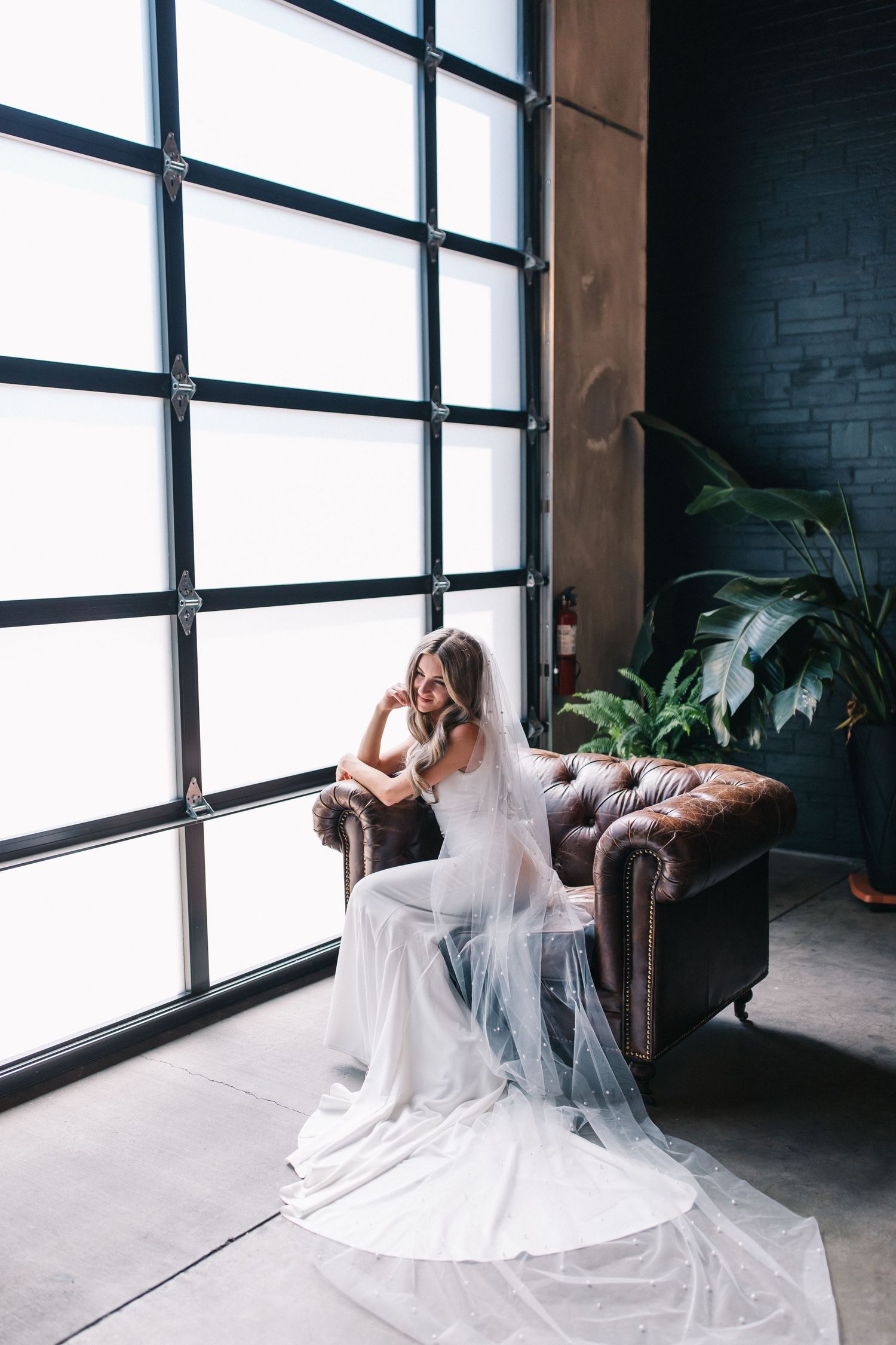 Wedding dress portrait by large industrial windows with flowing veil and leather chair.