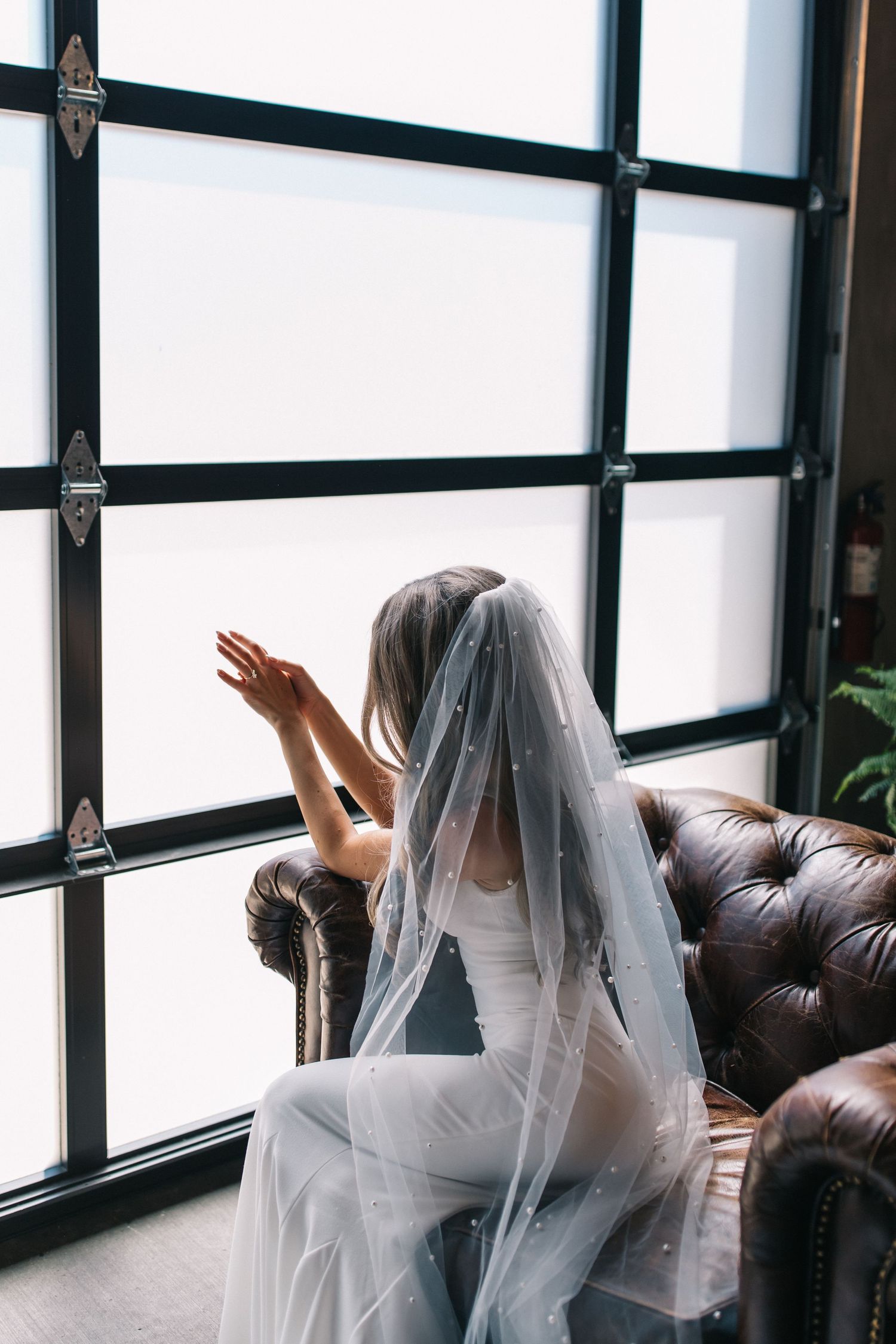 Artistic bridal photo with flowing veil against modern windows and leather furniture.