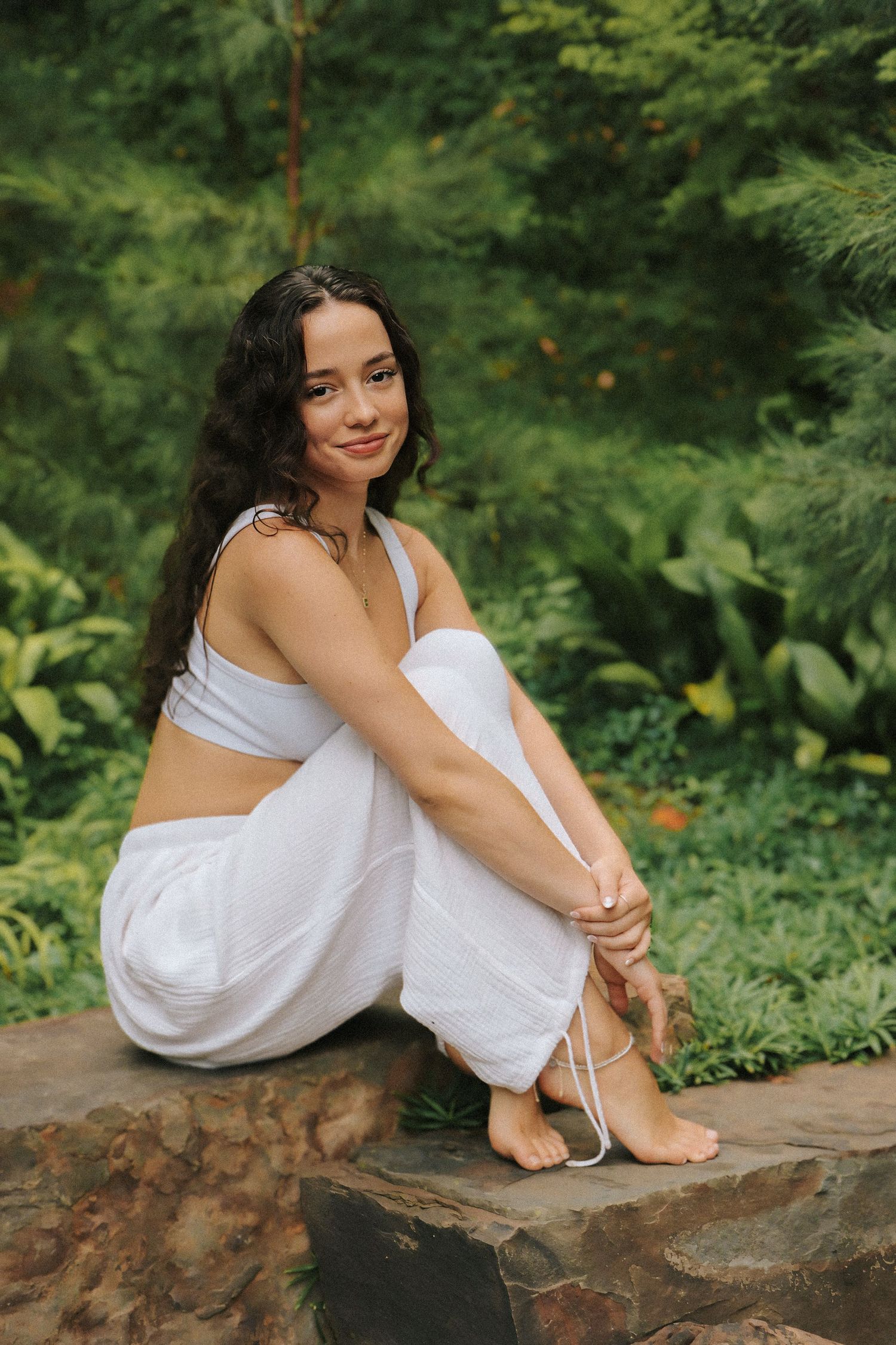 A woman in white loungewear sits peacefully on a stone ledge surrounded by lush greenery.