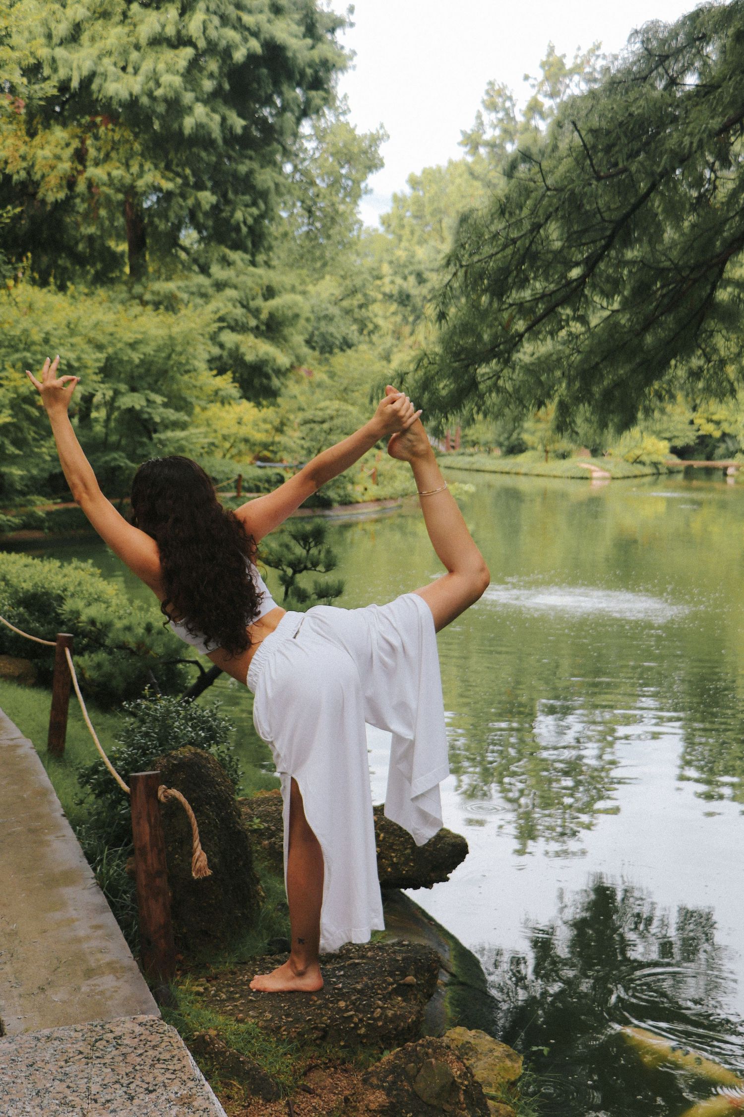 A dancer strikes a graceful pose by a serene pond surrounded by trees.