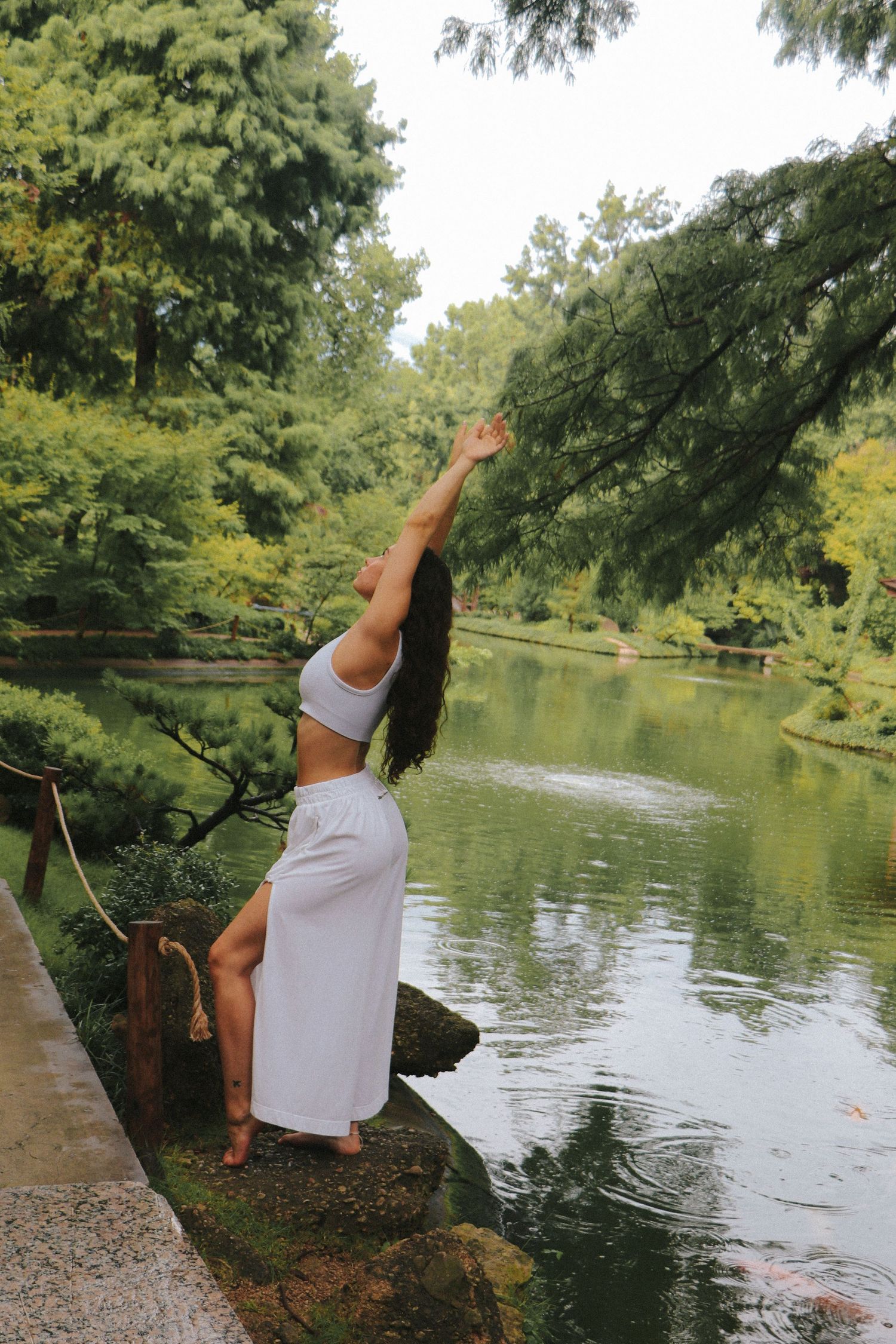 A person performs a ballet-inspired pose near a reflective pond in a garden setting.