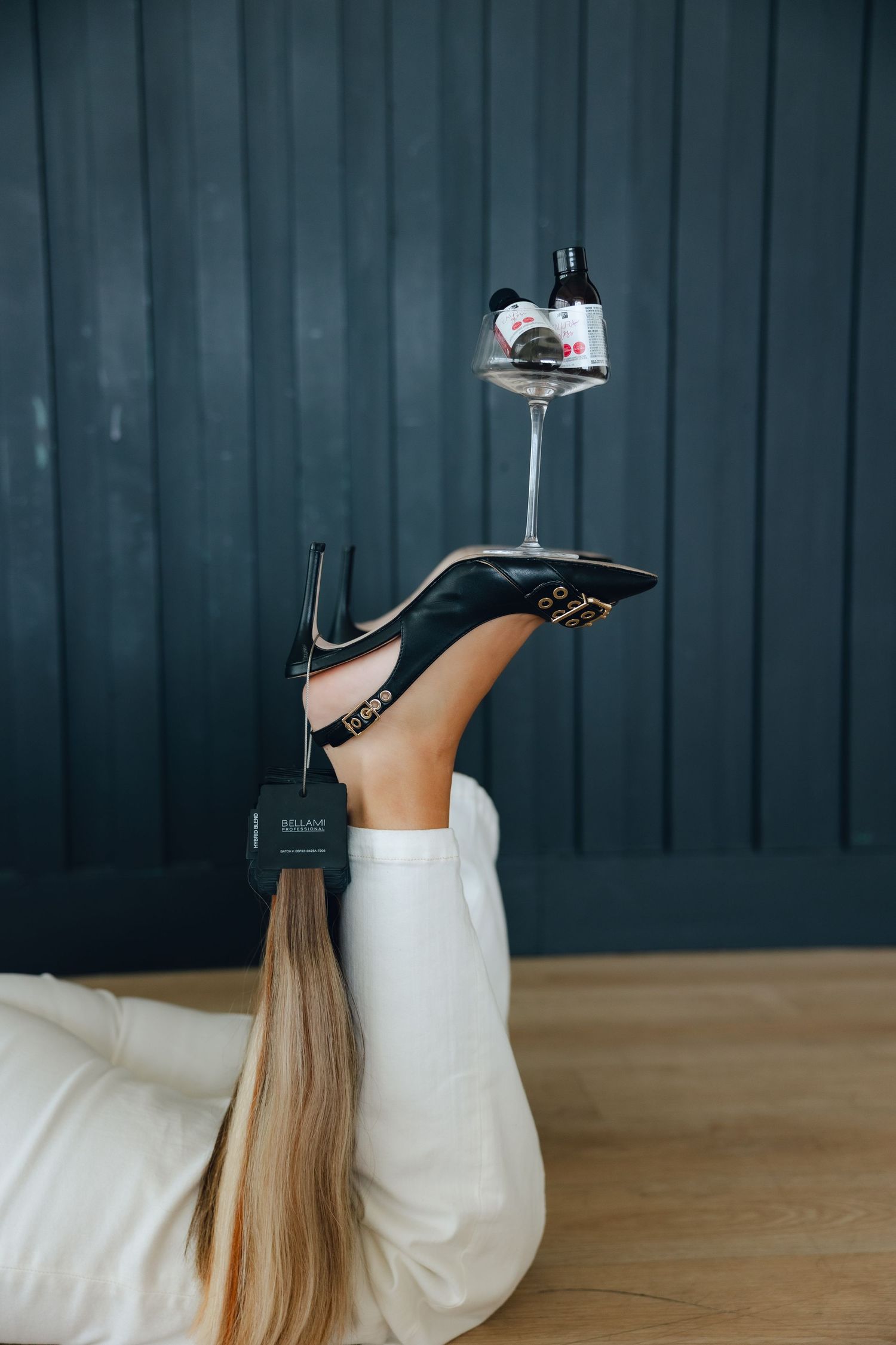A wine glass balanced on a black high heel shoe worn by someone lying on a wooden floor in white pants.