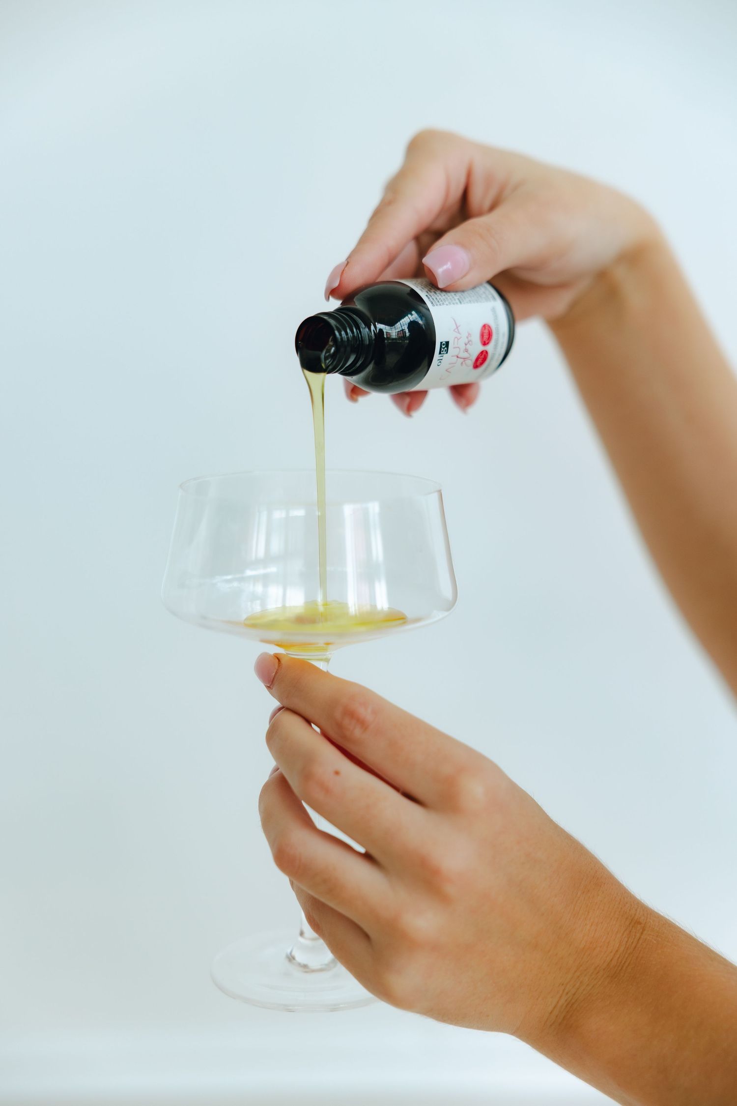 Hands pouring olive oil from a dark bottle into a small glass container against a white background.