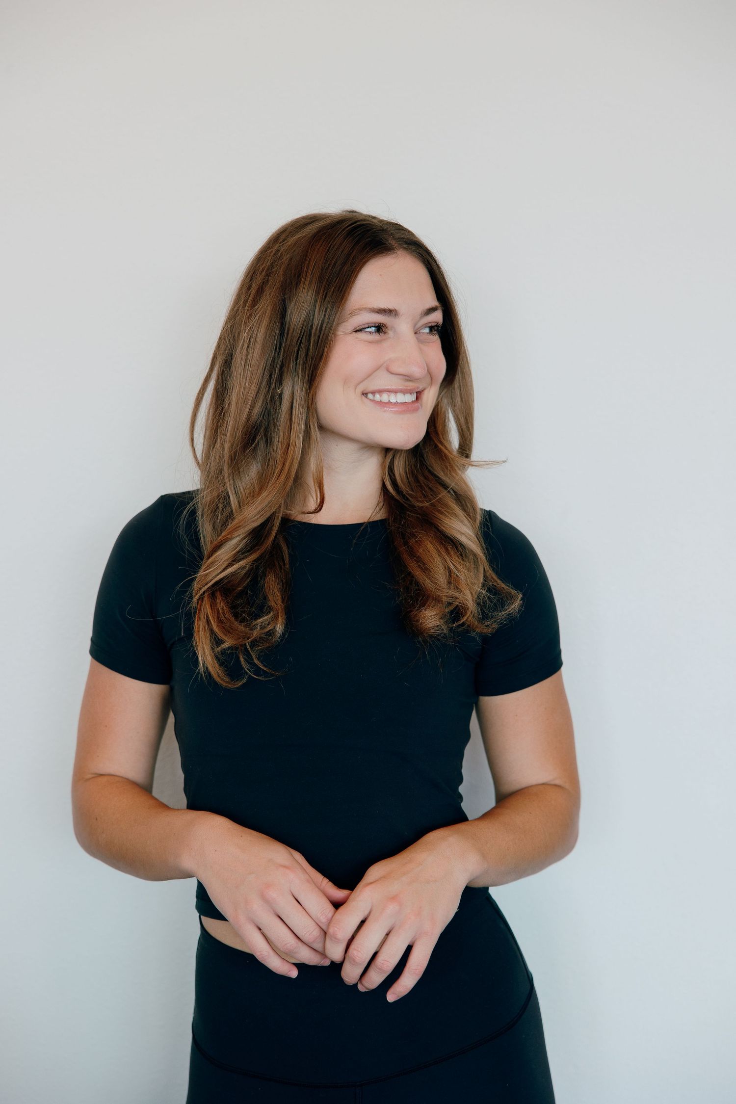 Professional headshot of a woman with wavy brown hair wearing a black dress against a light background.