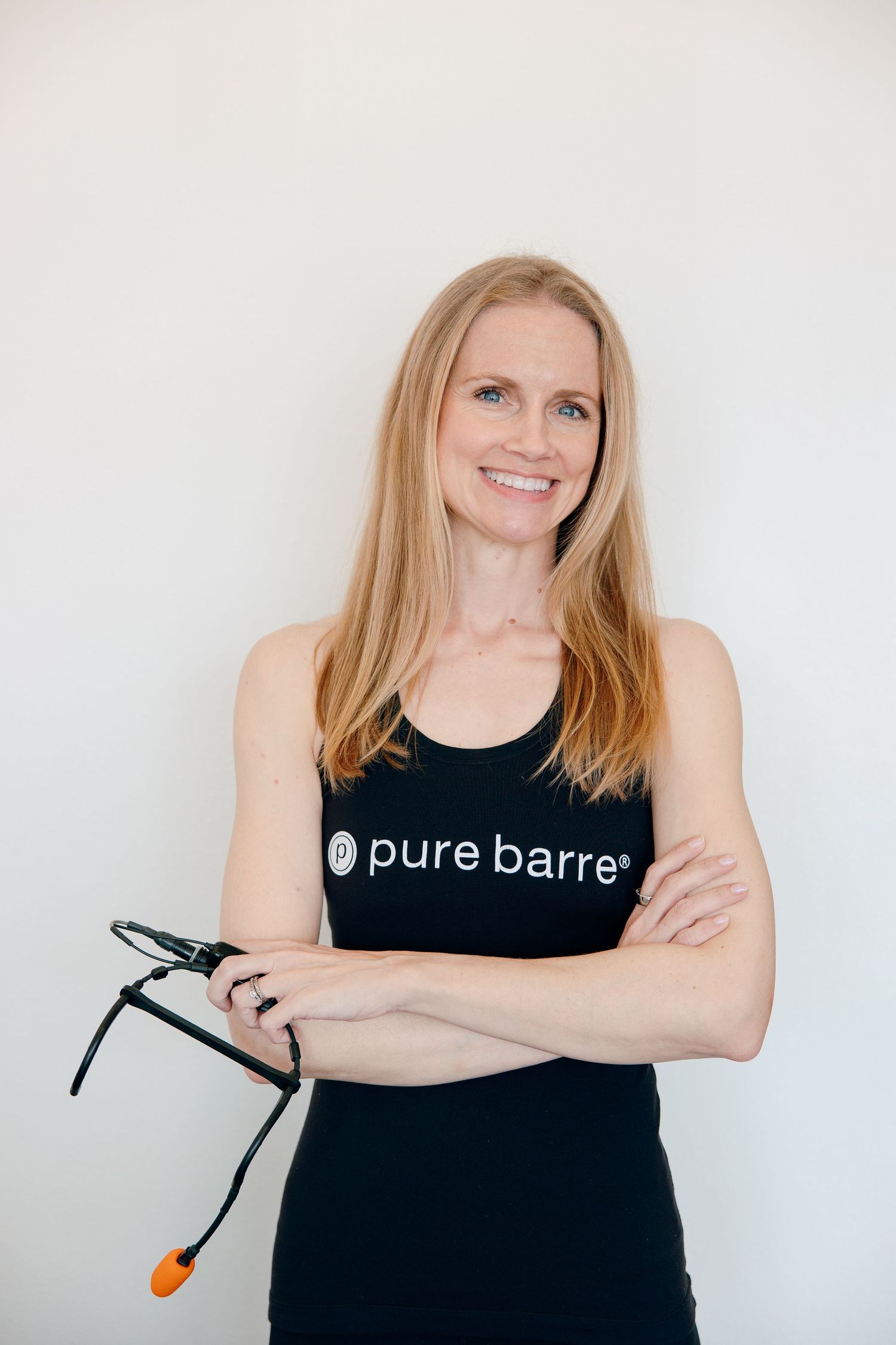 Fitness instructor wearing black Pure Barre tank top holds exercise band against white background.