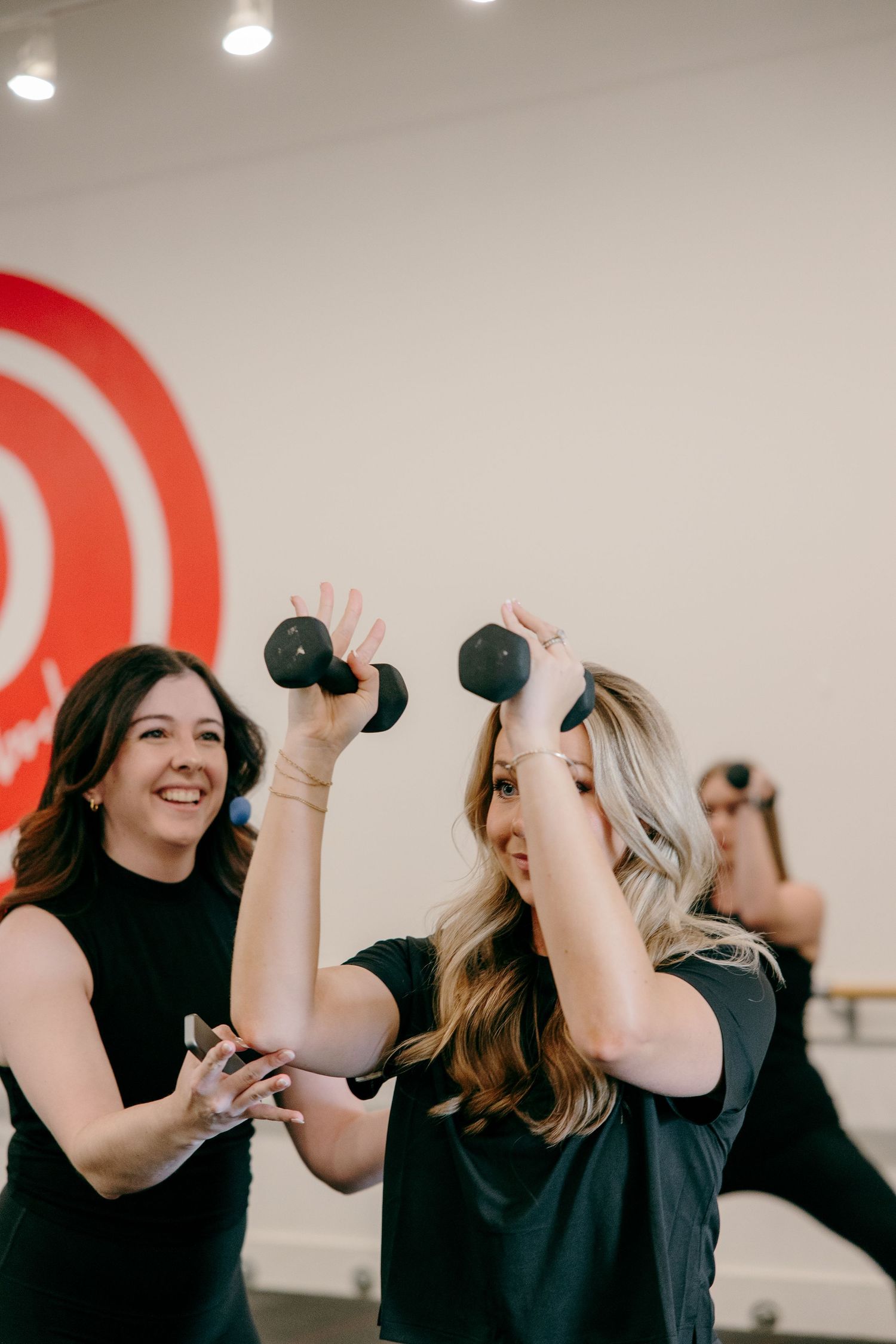 Two women in black workout clothes lift small dumbbells during a fitness class with a red target logo on the wall.