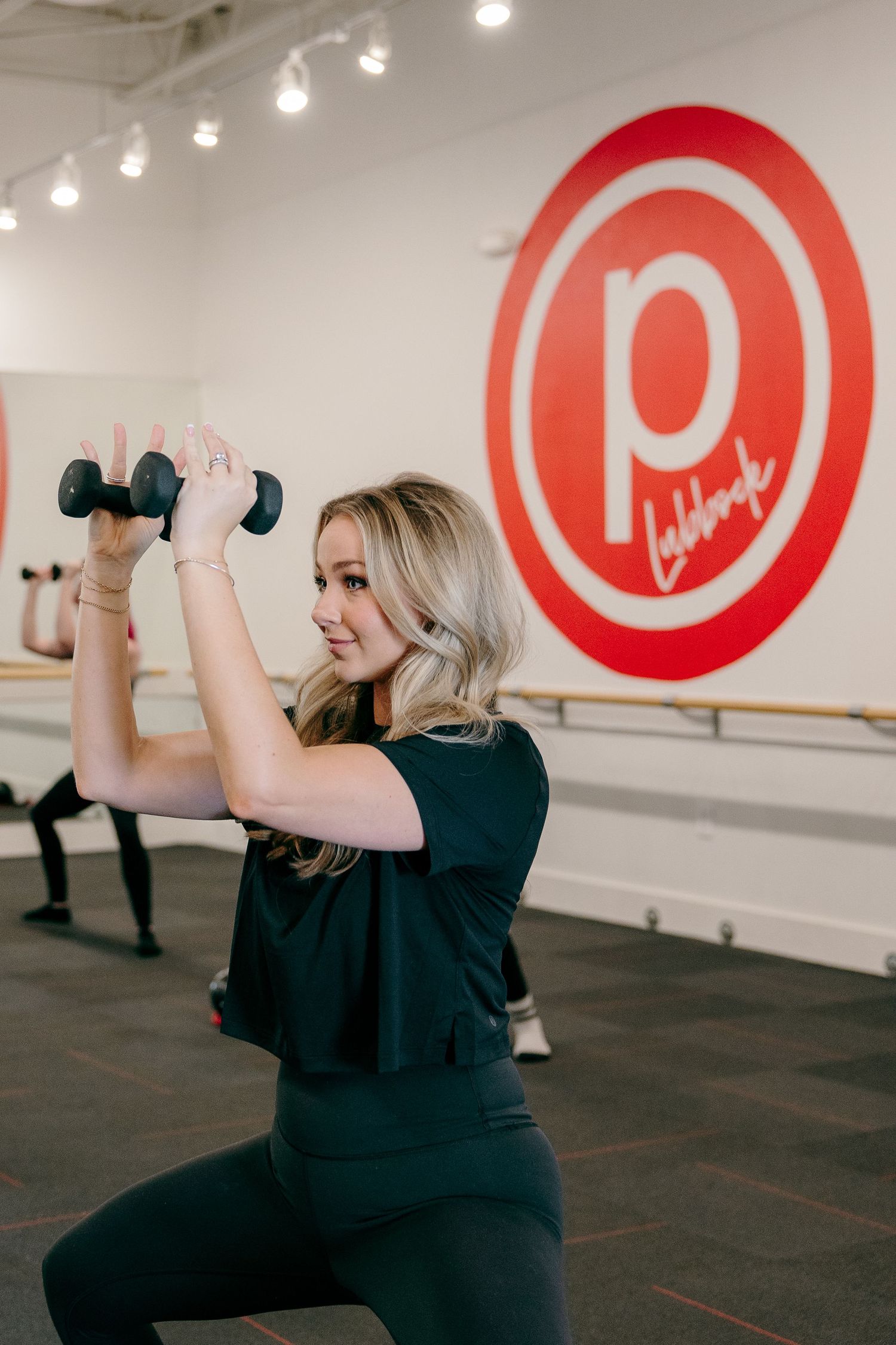 Person exercising with dumbbells in a Pure Barre fitness studio with red logo on white wall.