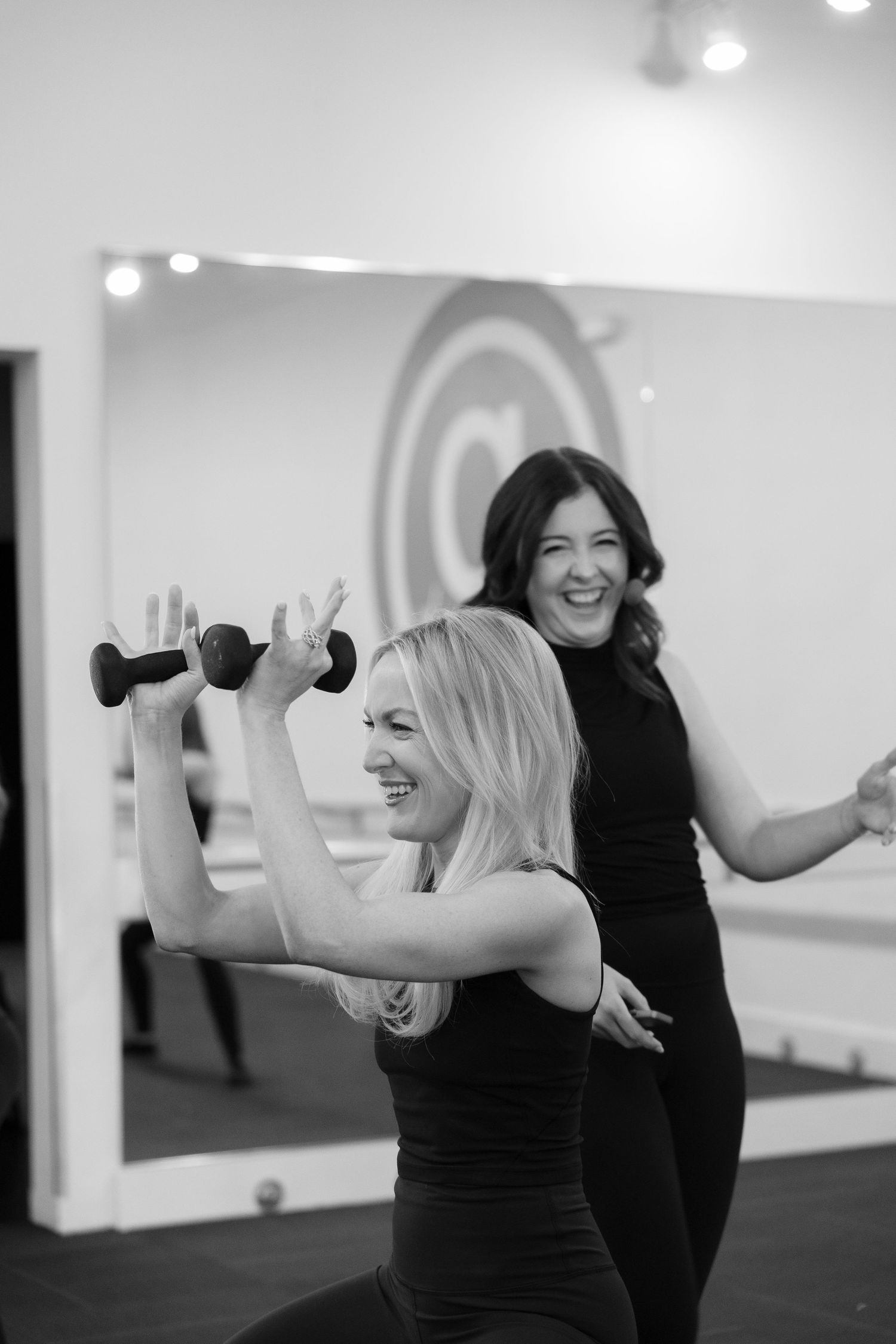 Two women in black workout attire laughing during a fitness class with one holding dumbbells.