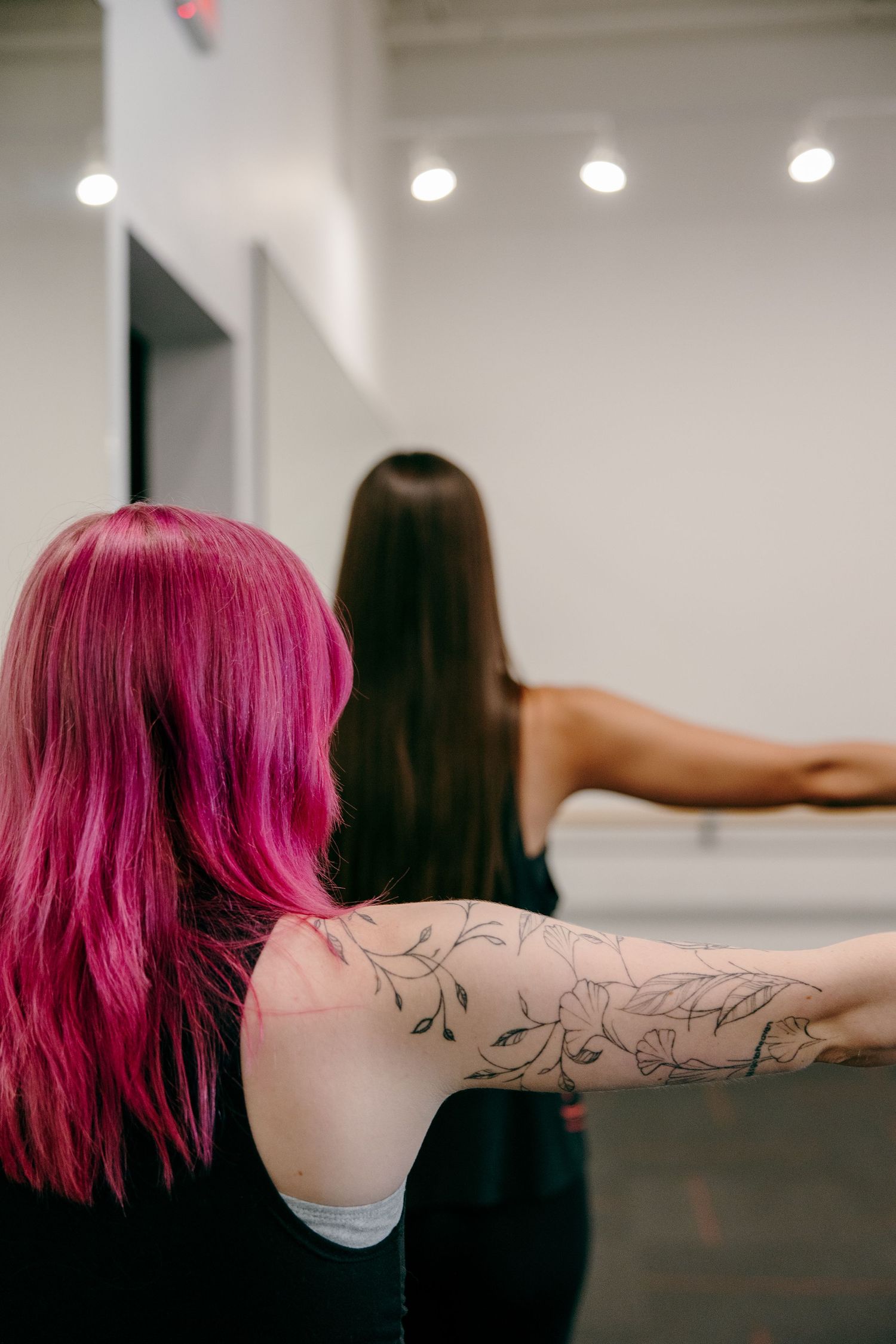 Two women stretching their arms during a workout class, with one sporting bright pink hair and visible tattoos.