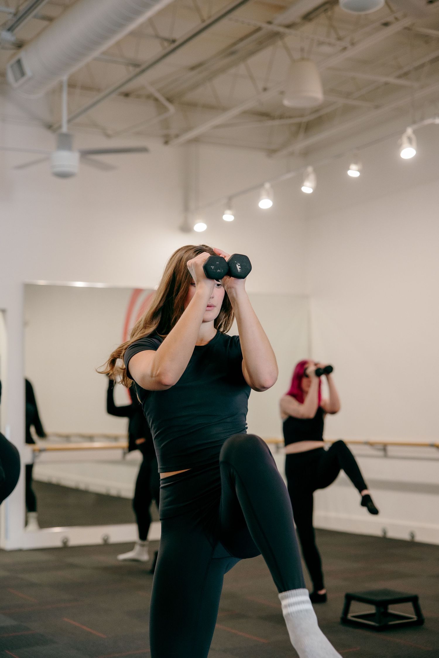 Athletic participant exercises with hand weights during a workout class in a studio with mirrors.