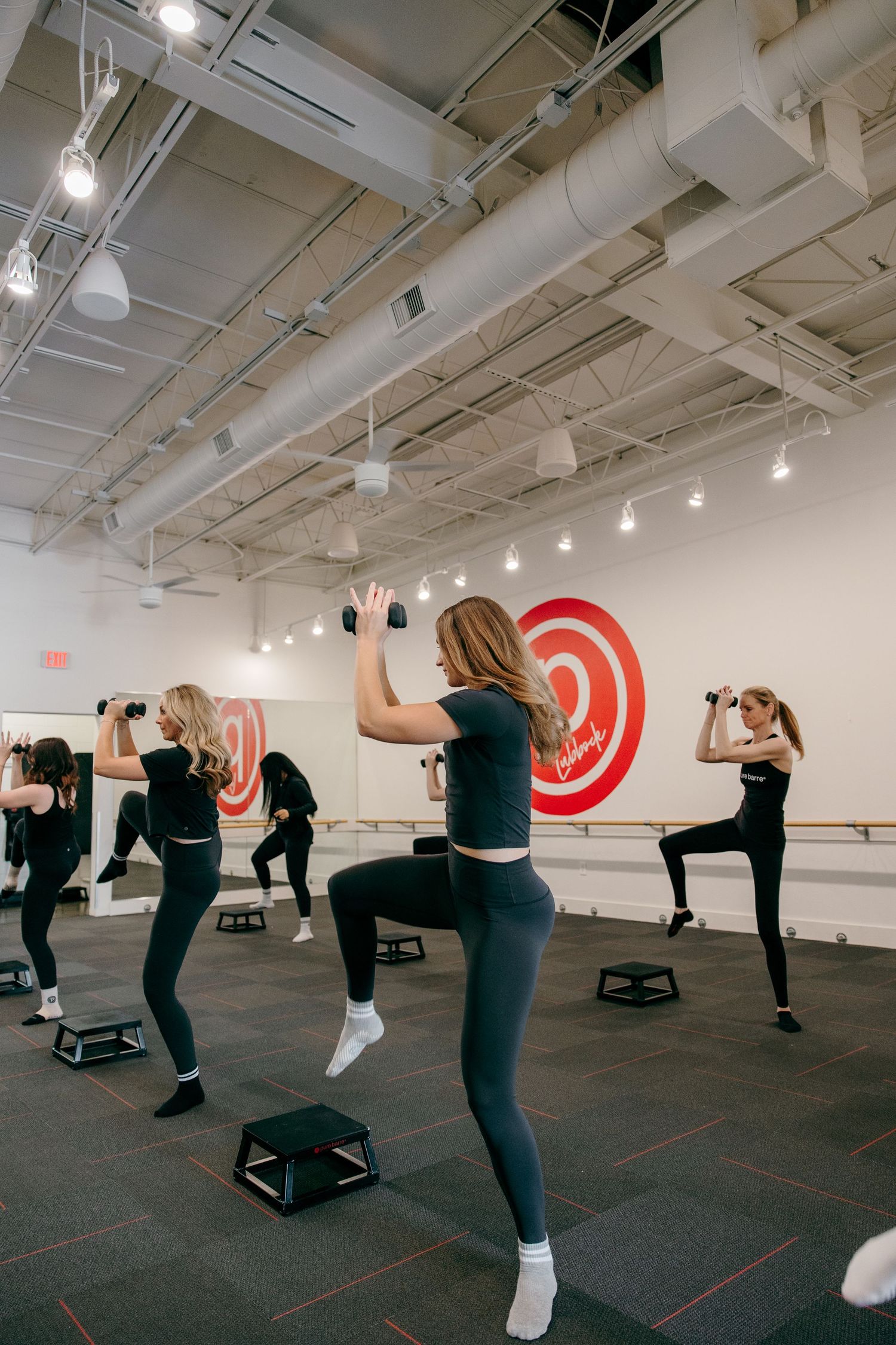 Group fitness class performs high kicks with step platforms in a studio with red target wall decor.