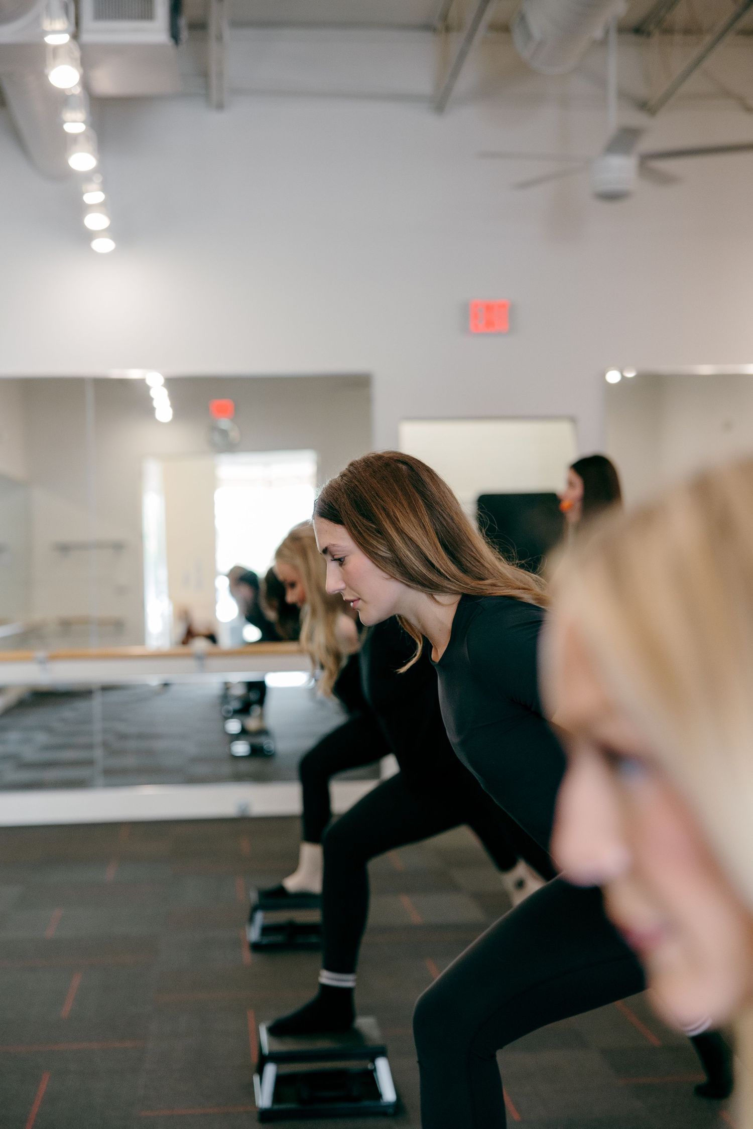 Fitness class participants doing step exercises in a mirrored studio with bright overhead lighting.