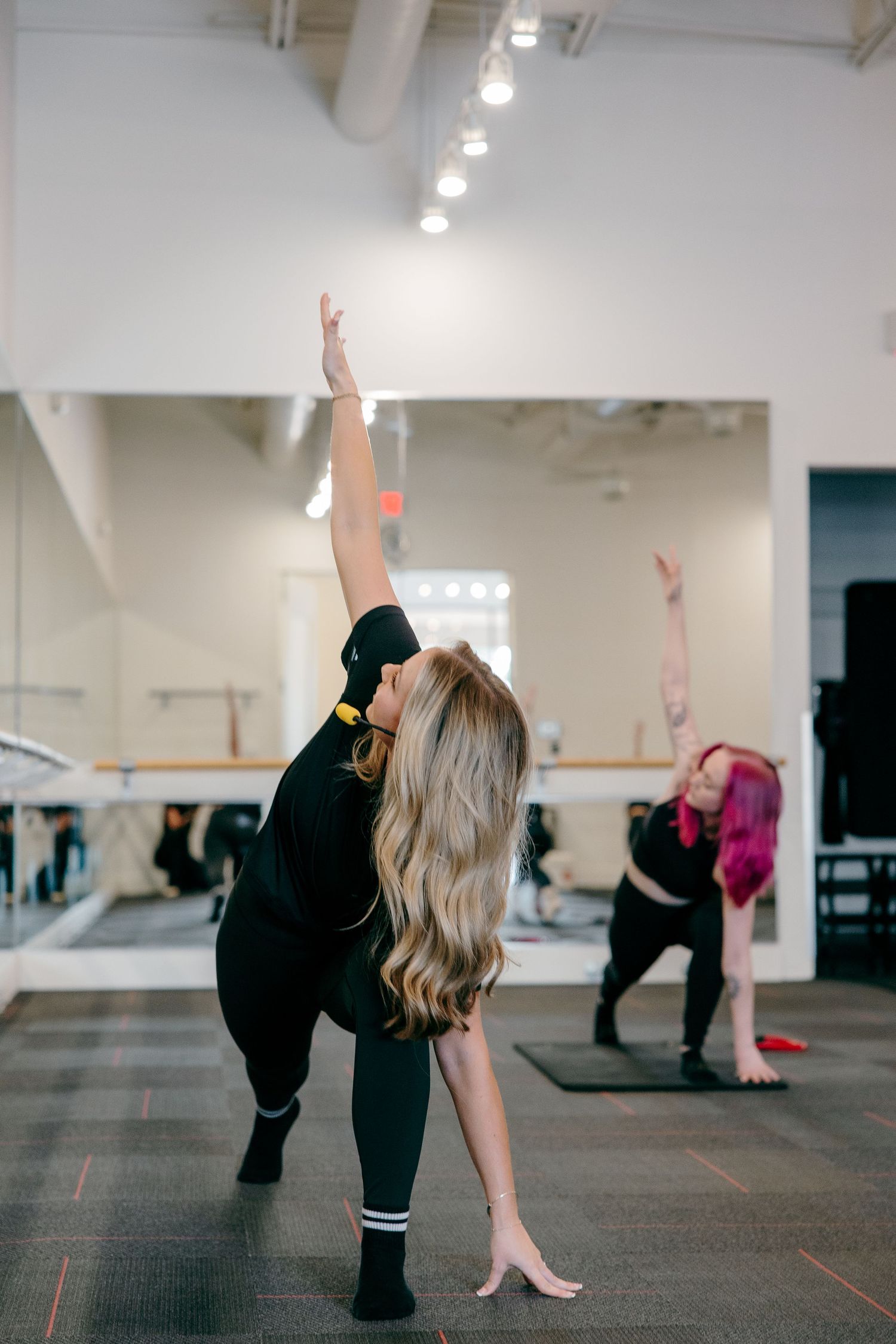 Students practicing dance movements in a mirrored fitness studio with overhead lighting and a red logo.