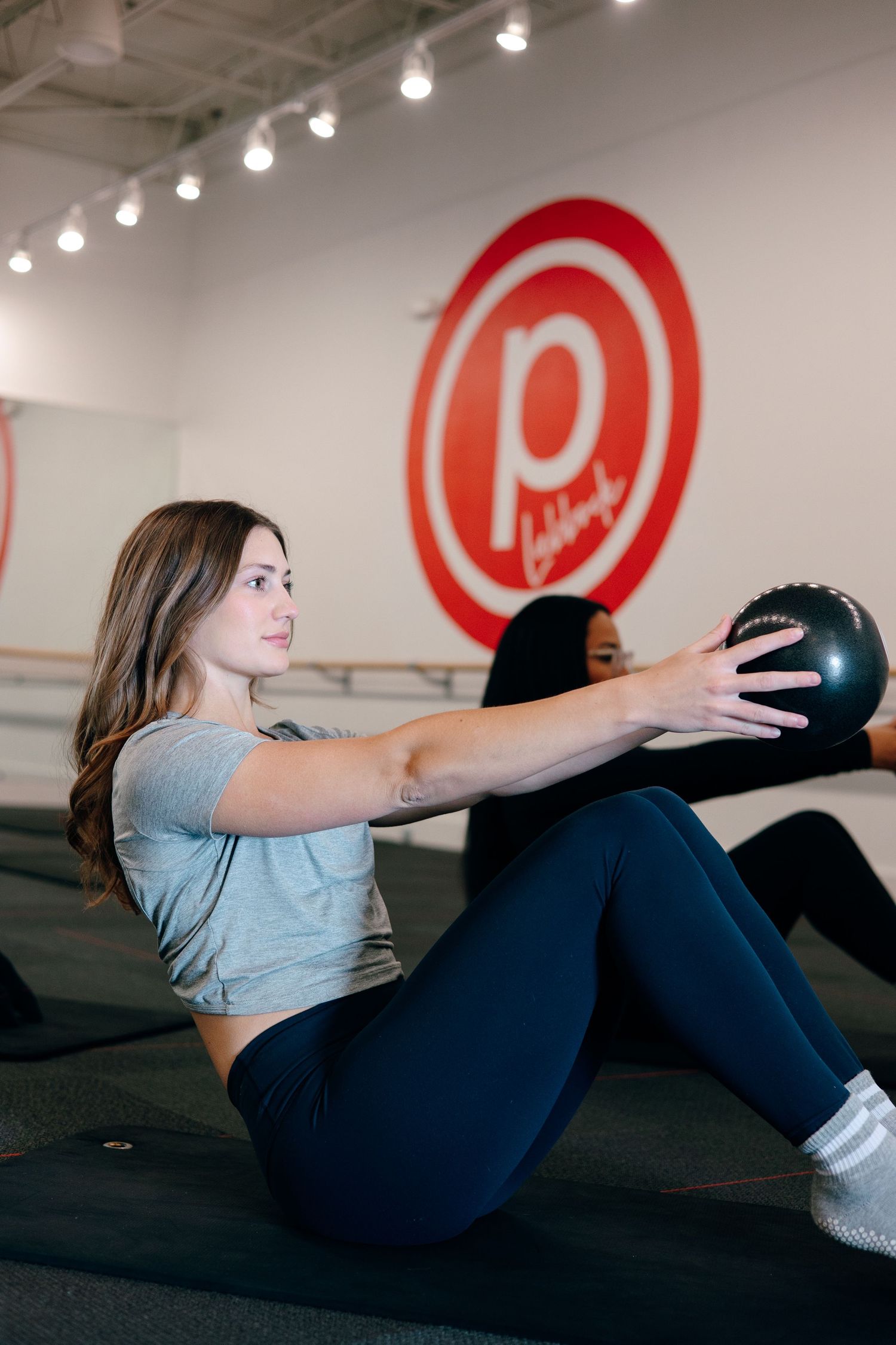 Participant performs core exercise with medicine ball in modern fitness studio featuring red circular branding.
