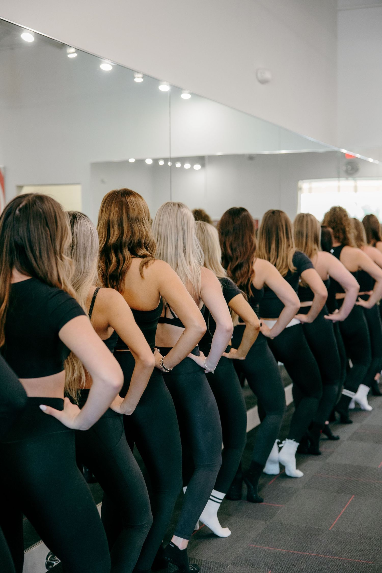Dancers in black outfits perform a synchronized line dance routine in a bright studio space with track lighting.