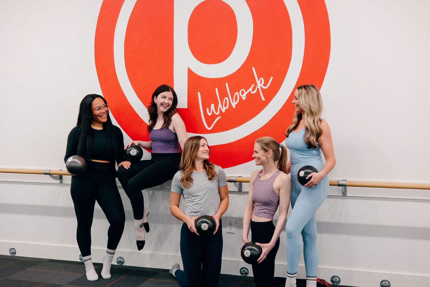 Group of workout participants posing with weights in front of red Pure Barre logo on studio wall.