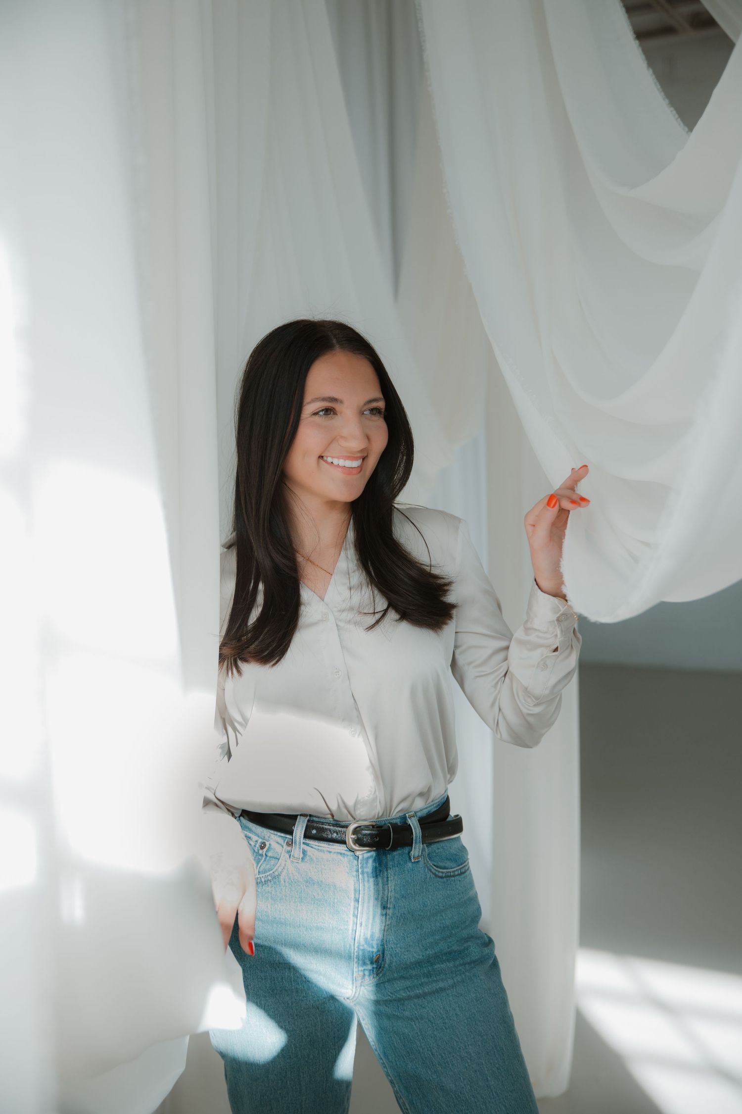 Woman in white blouse and jeans poses against white curtains in bright sunlit room.
