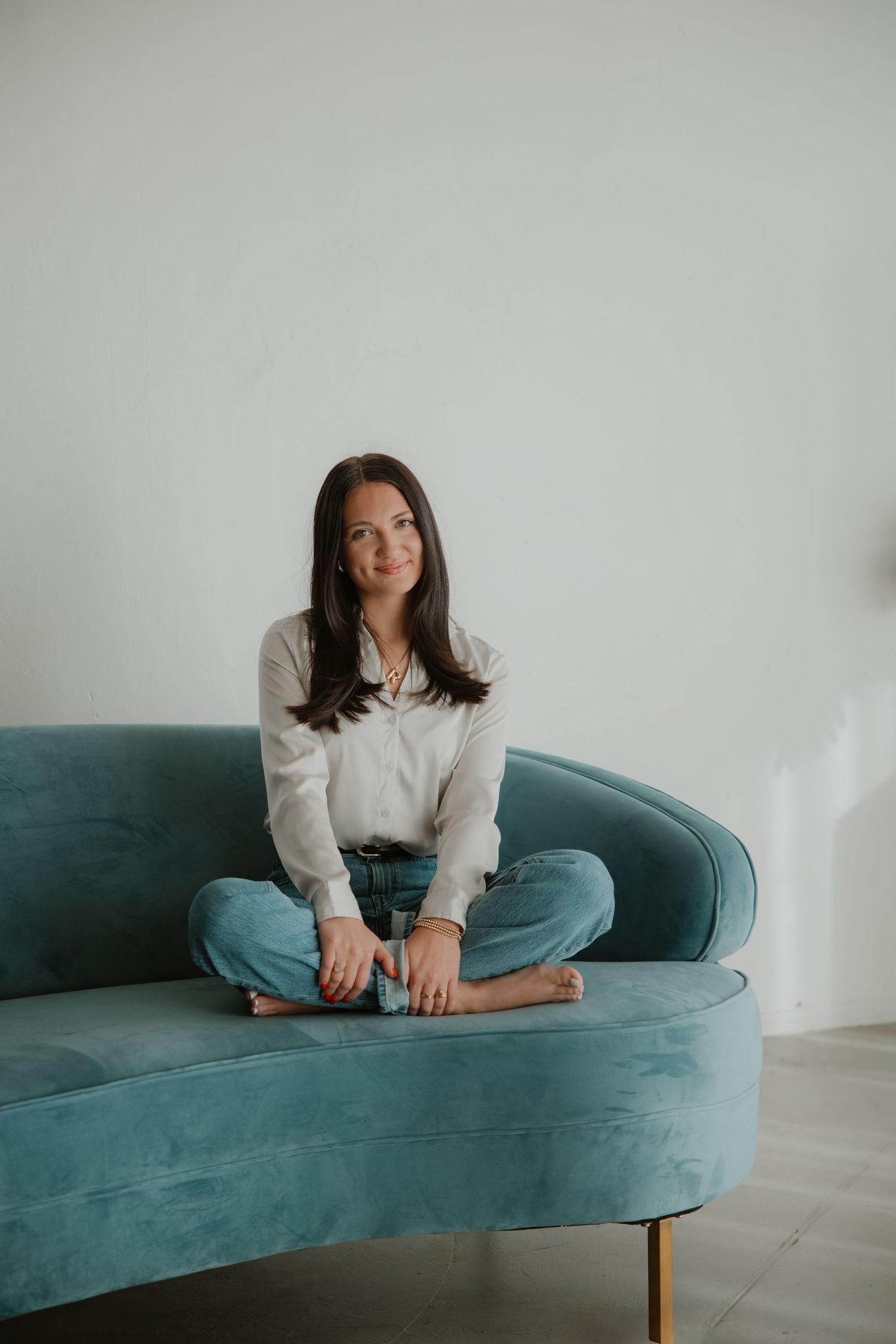Woman in casual attire sitting cross-legged on teal velvet couch against white wall.