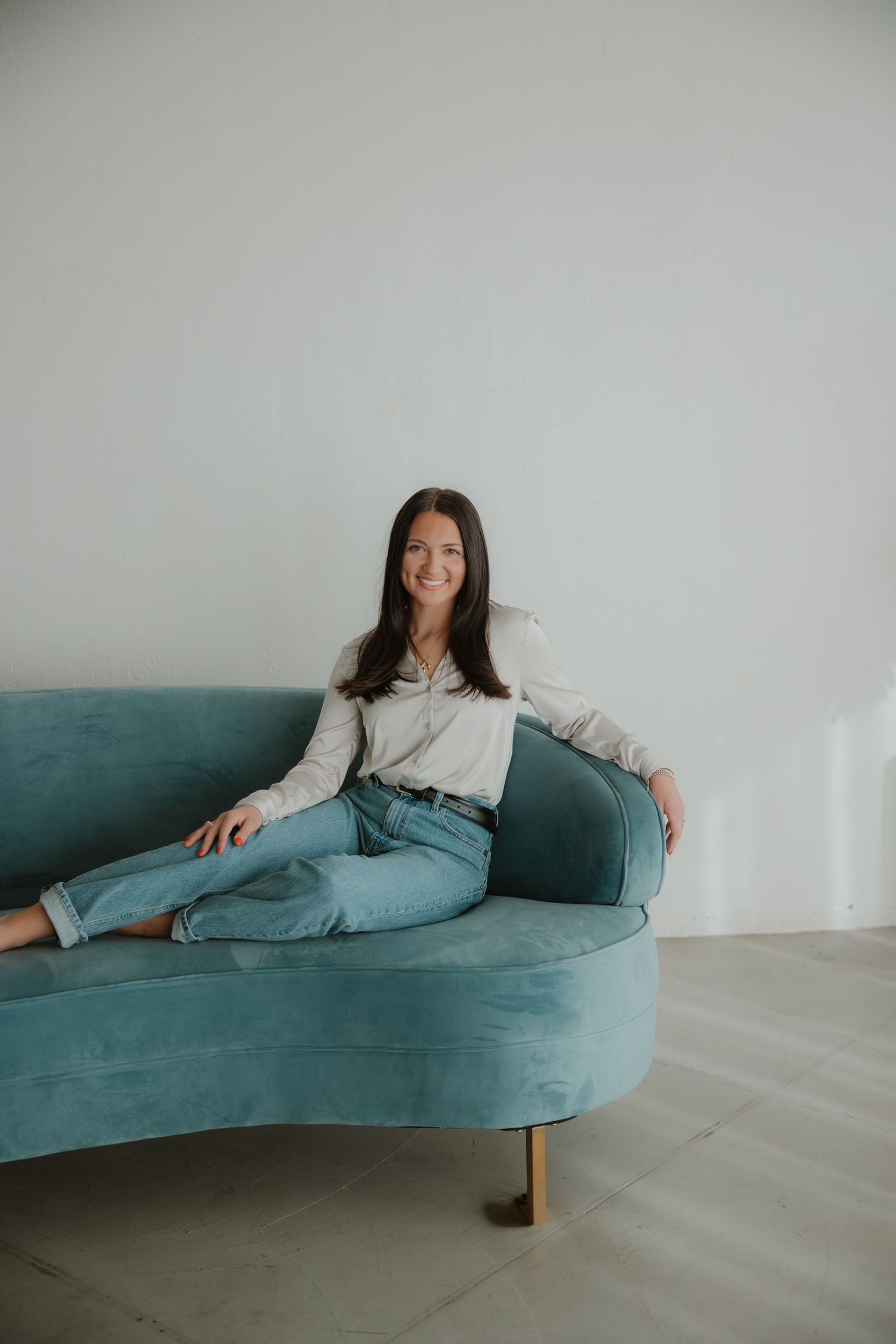 Woman relaxing on curved teal velvet sofa in minimalist room setting.