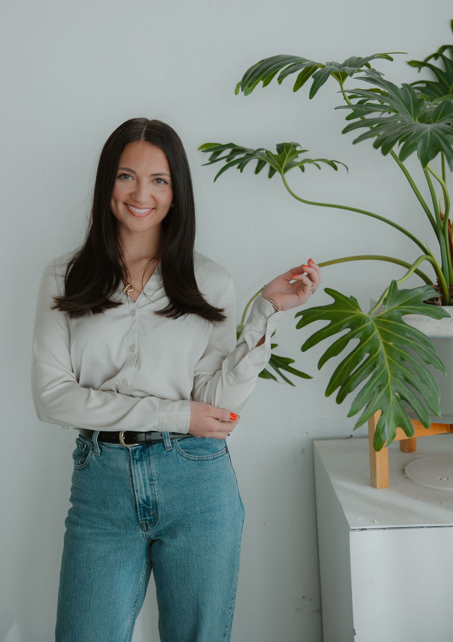 Woman in white shirt and jeans standing beside large monstera plant against white wall.