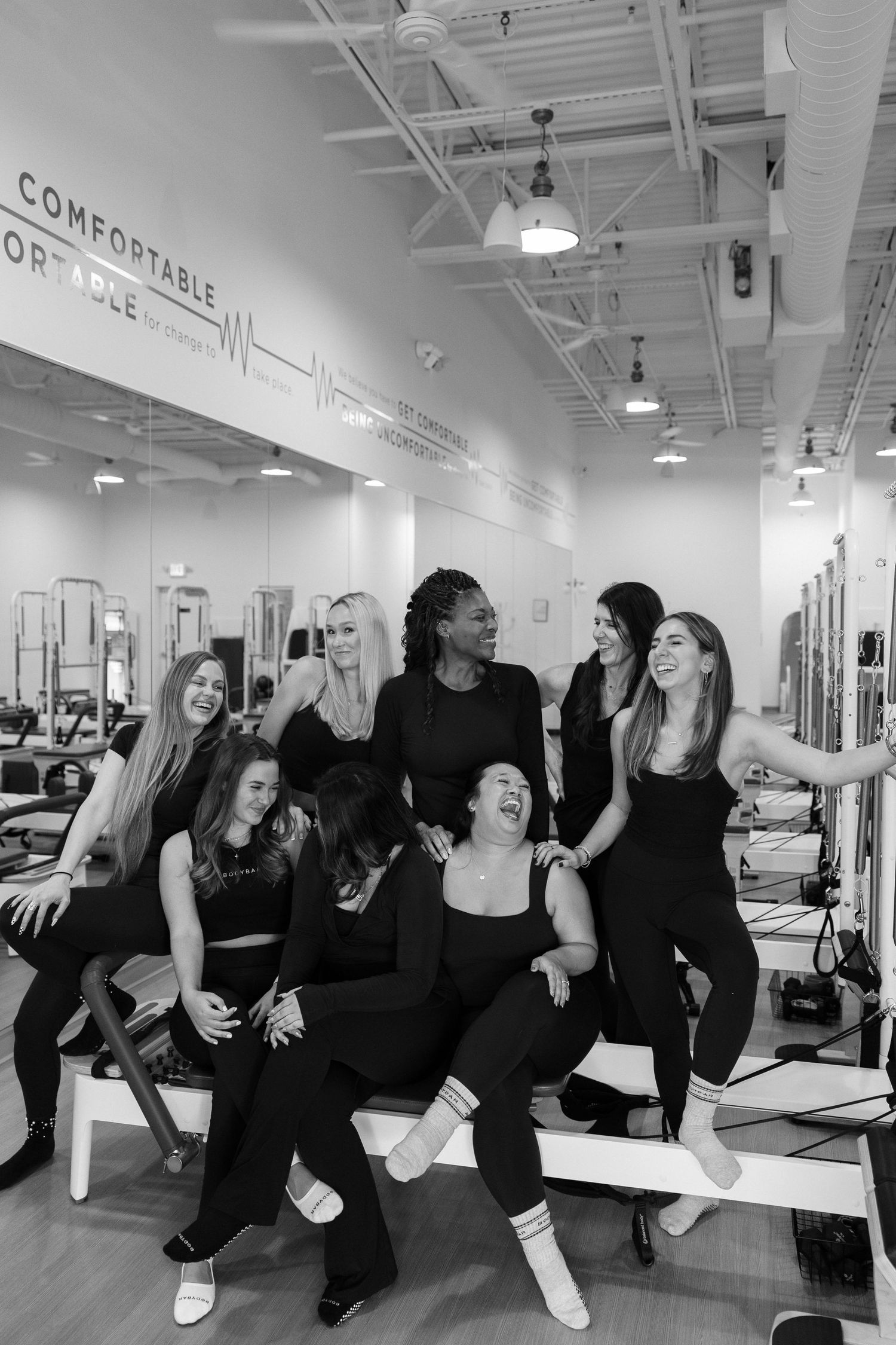 A group in black workout attire poses playfully together at a fitness studio with white walls and exercise equipment.