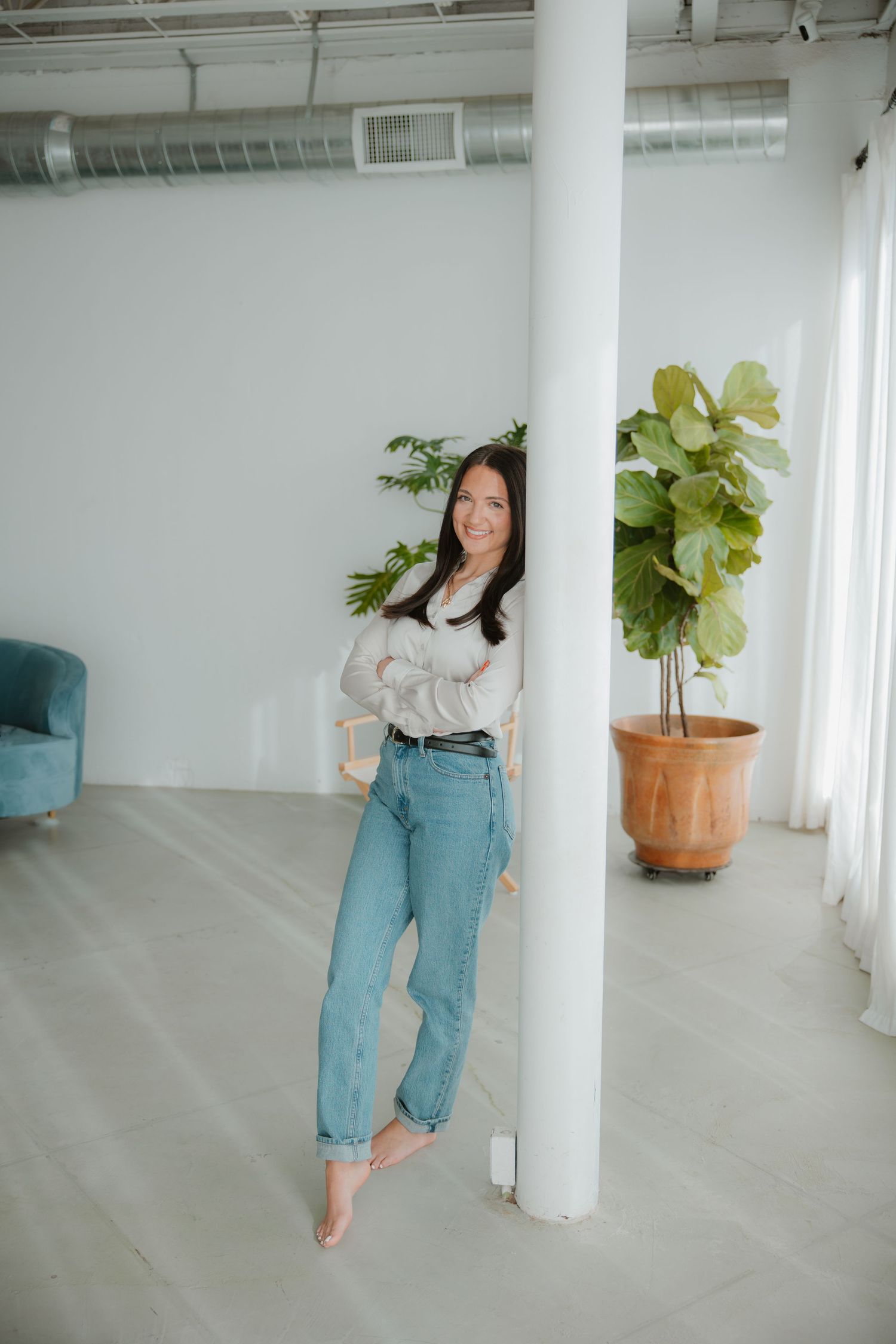 Woman in casual outfit posing barefoot by white column with indoor plant.