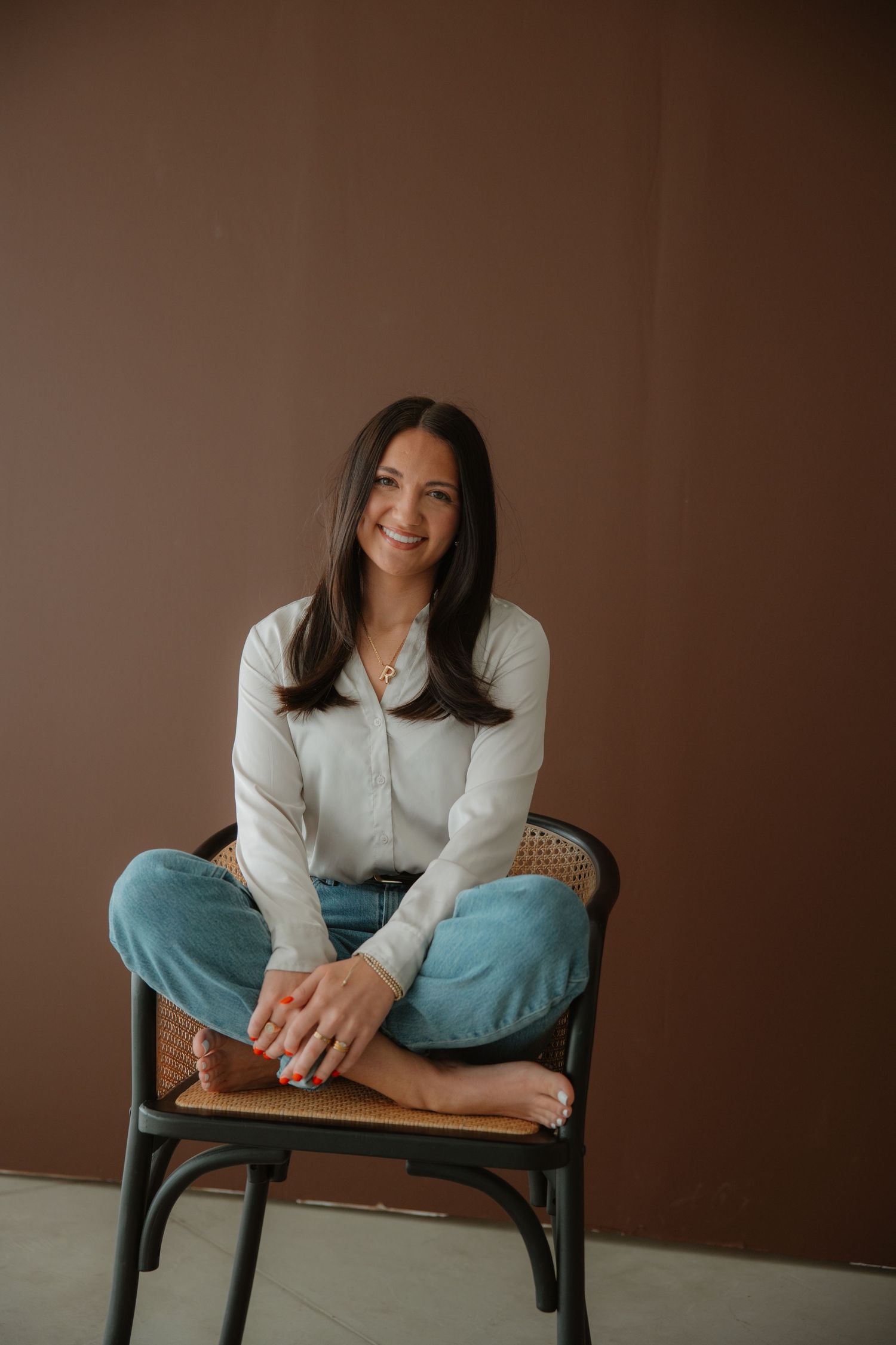 Woman in white shirt seated cross-legged on rattan chair against brown wall.