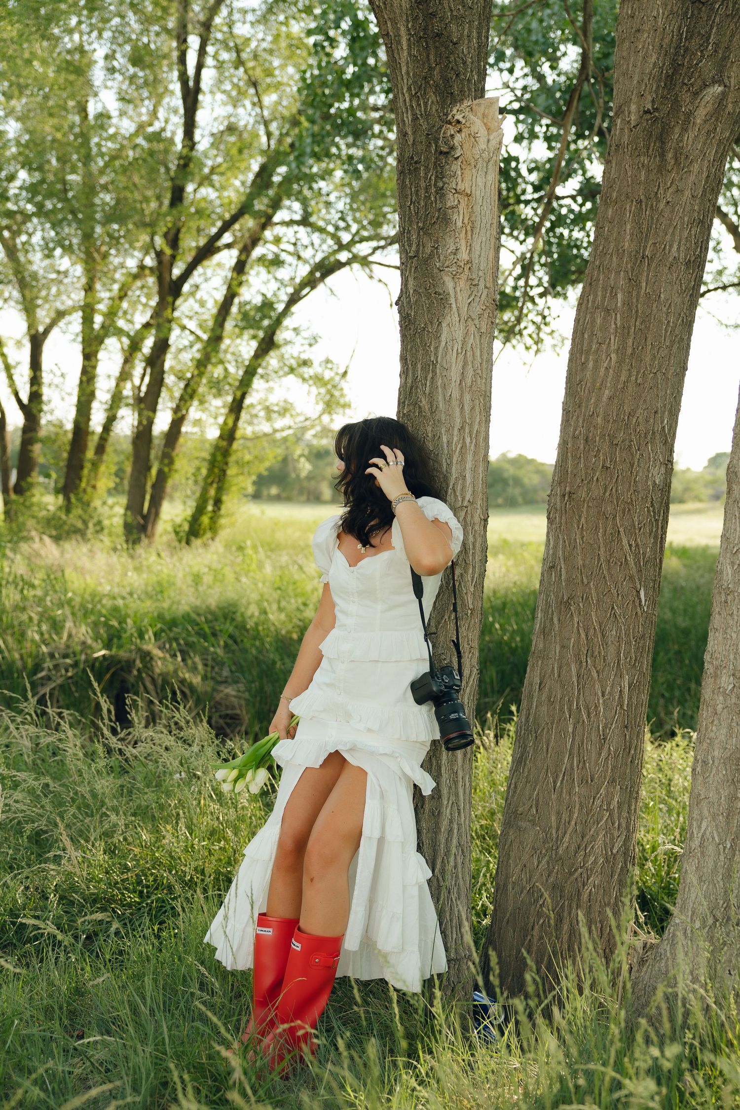 A woman in a white wedding dress poses playfully against a tree while wearing bright red rain boots in a nature setting.